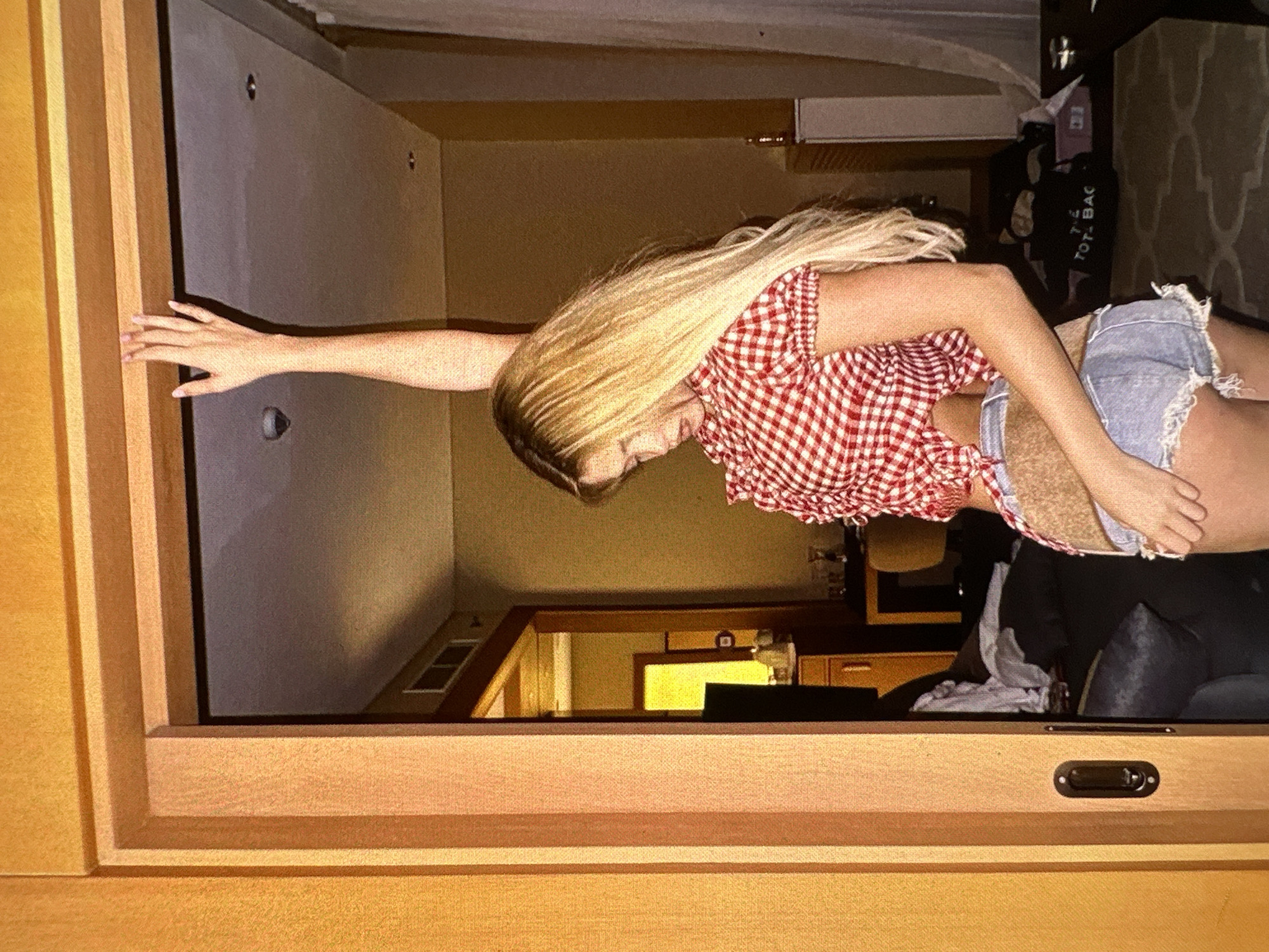 A woman stands in a room, framed by a wooden doorway. She has long blonde hair and is wearing a red and white checkered shirt and denim shorts. Her left arm is extended, reaching toward the left side of the frame. The room behind her includes a desk with a lamp, a couch, and other furniture. The location is Unknown, Unknown.
