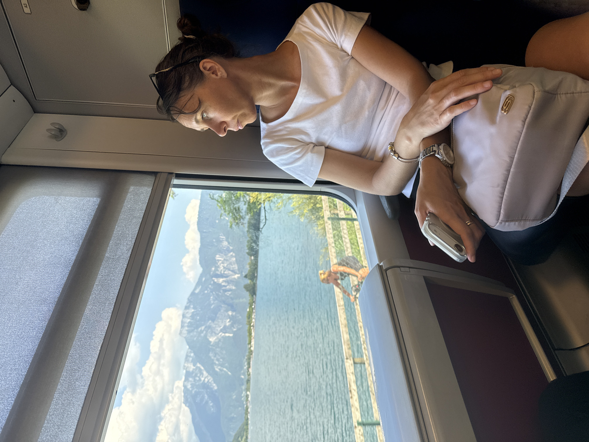 A woman is seated on a train, looking out the window at a picturesque mountain lake landscape. The train interior is visible, with a window and blinds on the left, and a seat and wall paneling on the right. The woman is wearing a white t-shirt and has her hair pulled back. She is holding a small silver object in her right hand and has a bag on her lap. Her left arm is resting on the bag, and she is wearing a watch on her left wrist. In the distance, through the window, a lake is visible with mountains and clouds in the background. A person on a bicycle is also seen near the lake. The image is taken from a slightly elevated angle, focusing on the woman and the view. The specific location of Pergine Valsugana, Italy, is not discernable from the image itself.