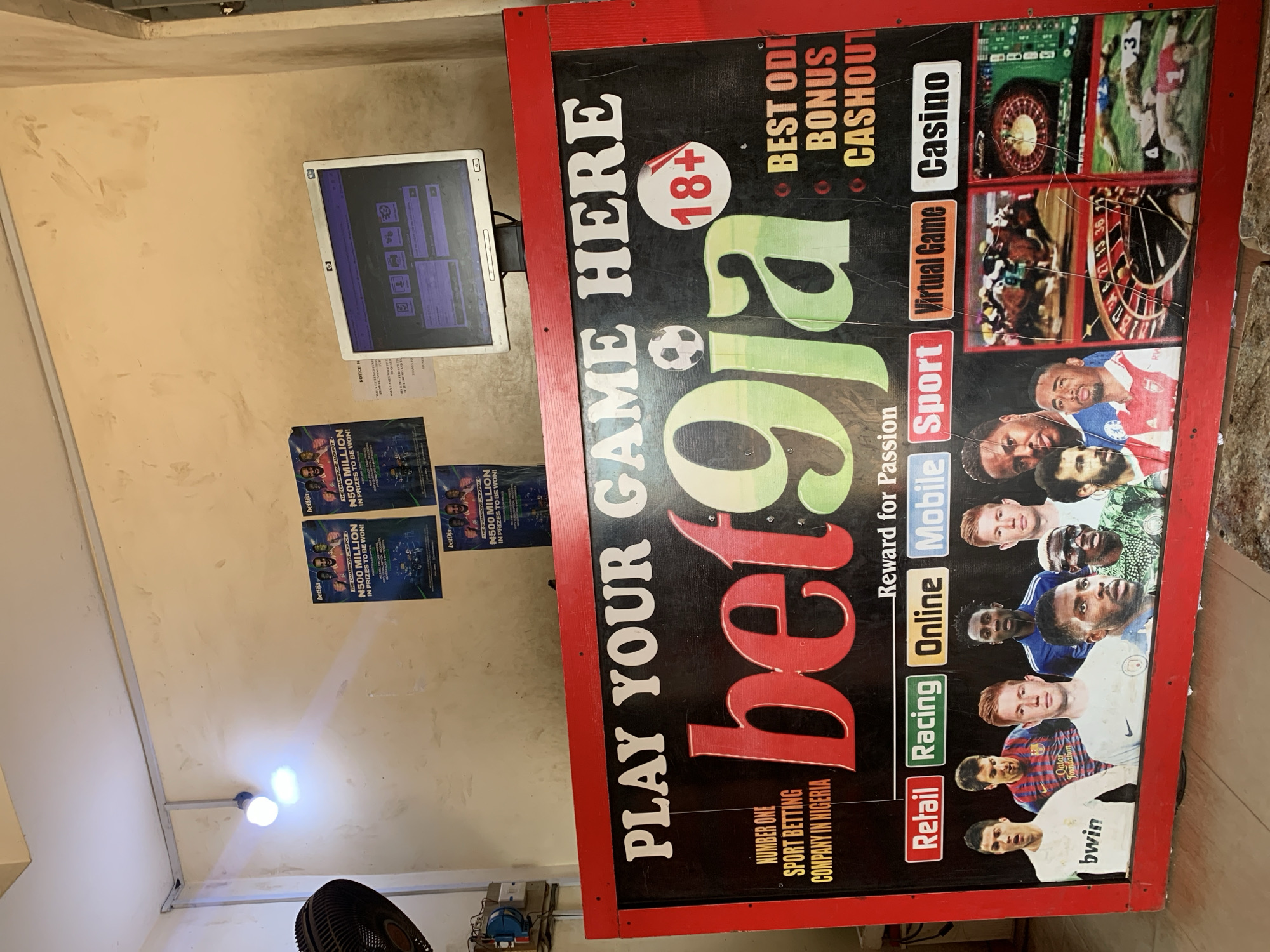 The image shows an interior space featuring a Bet9ja sports betting booth. The booth is identifiable by a large, red-framed sign with "PLAY YOUR GAME HERE" and the "bet9ja" logo prominently displayed. The sign also mentions "BEST ODDS BONUS CASHOUT" and lists various betting options like "Retail, Racing, Online, Mobile, Sport, Virtual Game, Casino." The sign features images of athletes. Below the athletes are football player images showing logos of "Bwin" and "Qatar Foundation". There is an HP computer monitor mounted on the wall next to the sign. Below the monitor are three Bet9ja posters promoting a N500 Million prize. The specific location within Adankolo, Nigeria, is not evident.