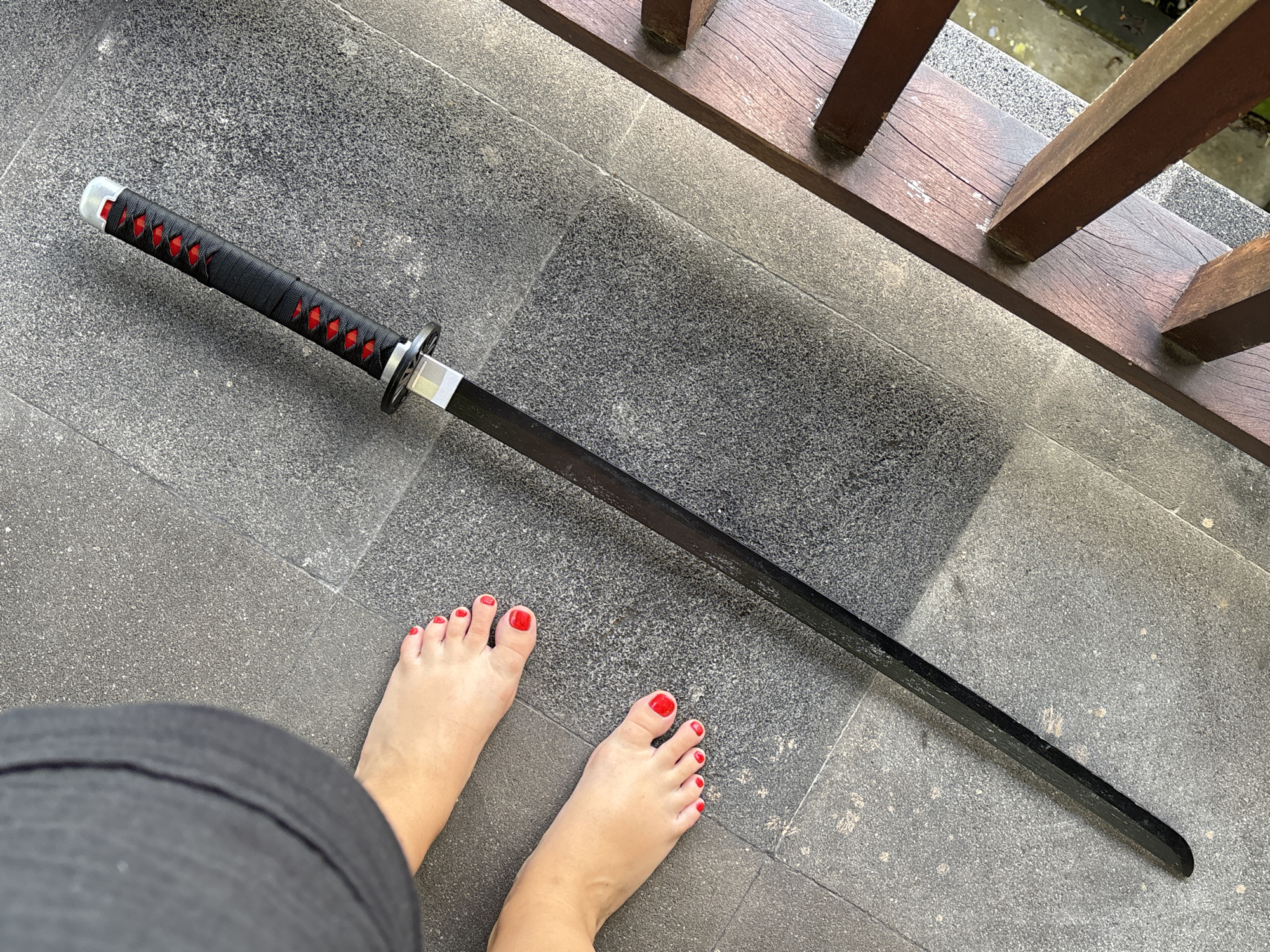 A full katana sword is lying on grey stone tiles next to a wooden railing. The sword has a black handle with red accents and a black blade. Near the sword are two human feet with red nail polish. The flooring appears to be outdoors, on the porch or a terrace, located in Kuta Selatan, Indonesia.