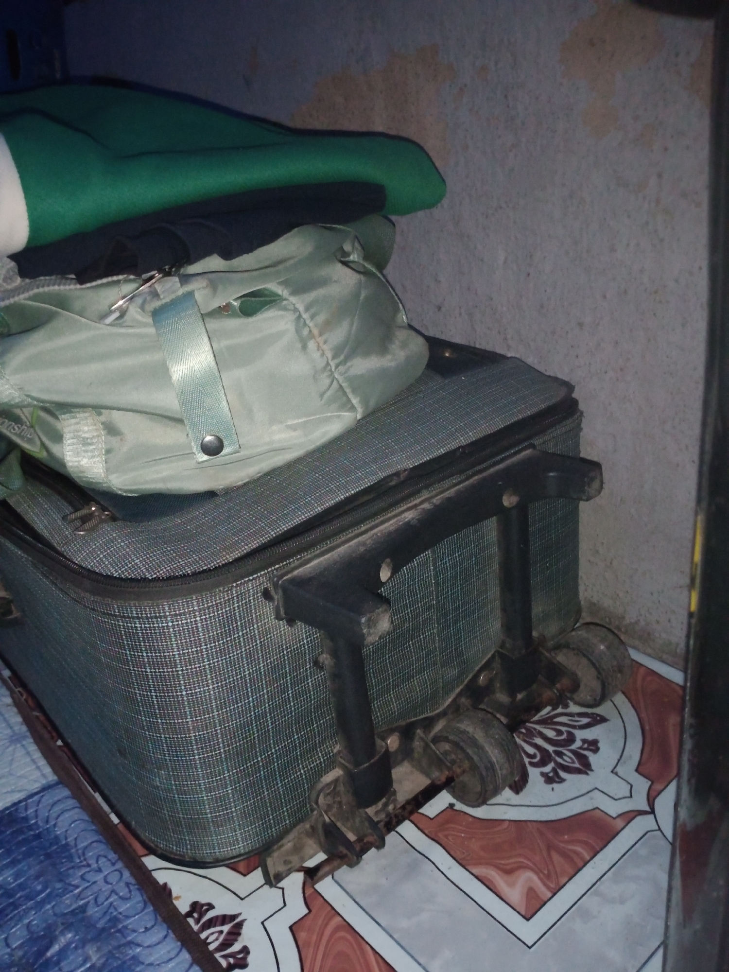 The image shows a stacked set of luggage against a textured, light-colored wall. The luggage consists of a green duffel bag placed on top of a larger, grey checkered suitcase with black wheels and a retractable handle. A dark blue item and a green blanket are partially visible on top of the duffel bag. The floor beneath the luggage has a tiled pattern featuring geometric and floral designs in blue, white, and brown. The overall scene appears to be indoors, likely in a storage area or bedroom. No people or specific locations within Jalingo, Nigeria are identifiable from the image.
