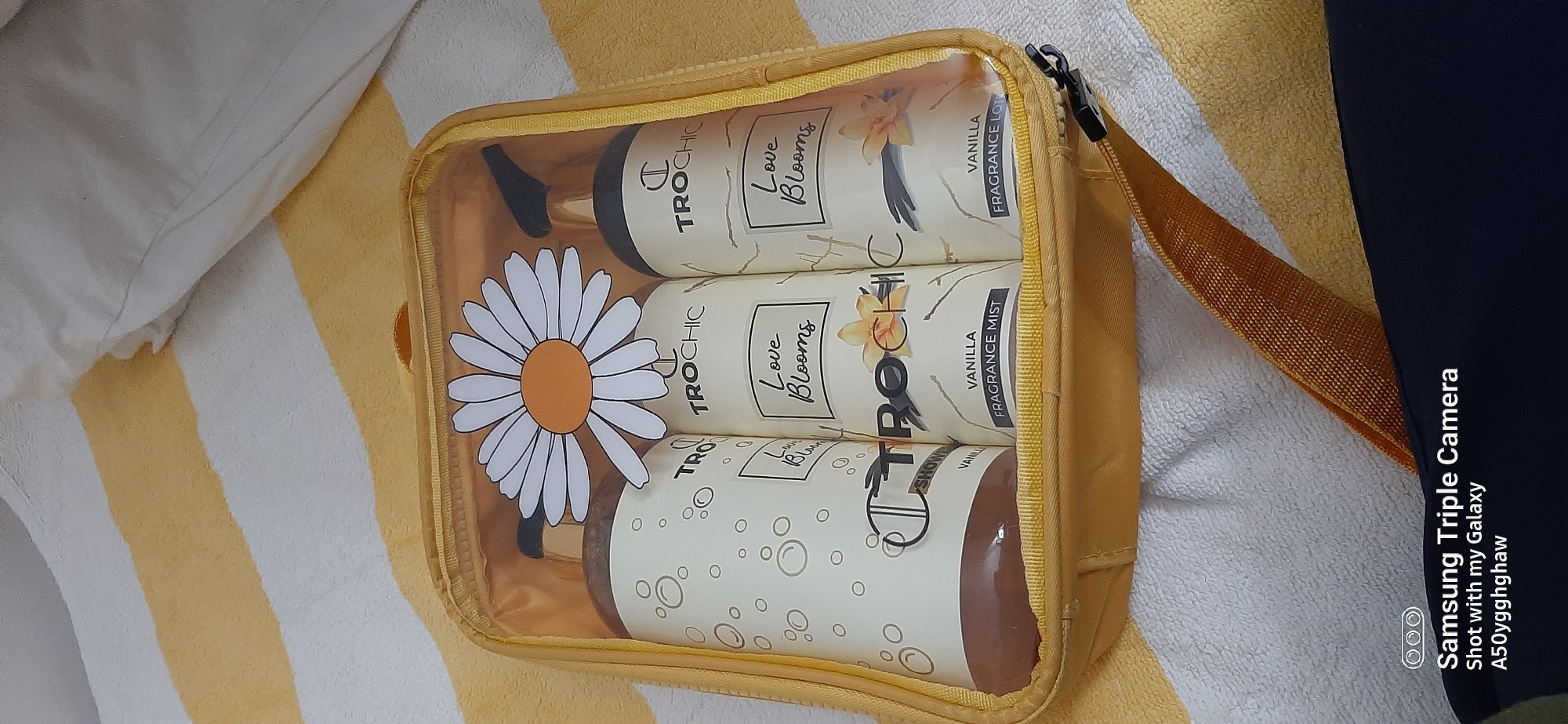 The image shows a close-up, eye-level, medium shot of a yellow cosmetic bag filled with three bottles of "Love Blooms" by TROCHIC. The bag is resting on a striped yellow and white towel. The bottles are arranged side-by-side and are partially visible through the clear plastic of the bag. The bottle on the far left is a shower gel, indicated by its brown liquid and the label "VANILLA SHOWER GEL". The middle bottle is a fragrance mist, labeled "VANILLA FRAGRANCE MIST". The bottle on the right is a body lotion, labeled "VANILLA FRAGRANCE LOTION". A daisy graphic is attached to the front of the cosmetic bag. The background consists of a white pillow and the striped towel. The overall impression is one of a product display, likely for sale or personal use. There are no people or animals visible in the image. The image was captured with a Samsung Triple Camera on a Galaxy A50. There is no information within the image that indicates the location in Dubai, United Arab Emirates.