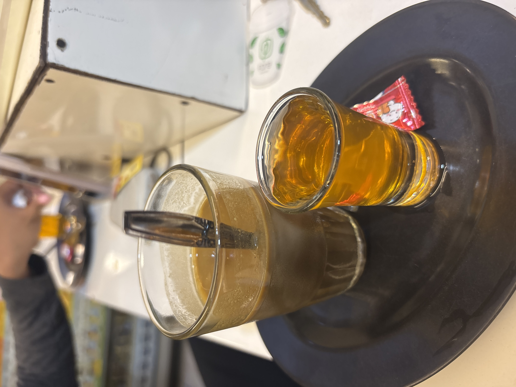 The image depicts two glasses on a dark plate. One glass contains a light-colored beverage with a spoon in it, while the other glass holds a yellow liquid. A packet lies next to the yellow liquid. The scene is in North Jakarta, Indonesia.