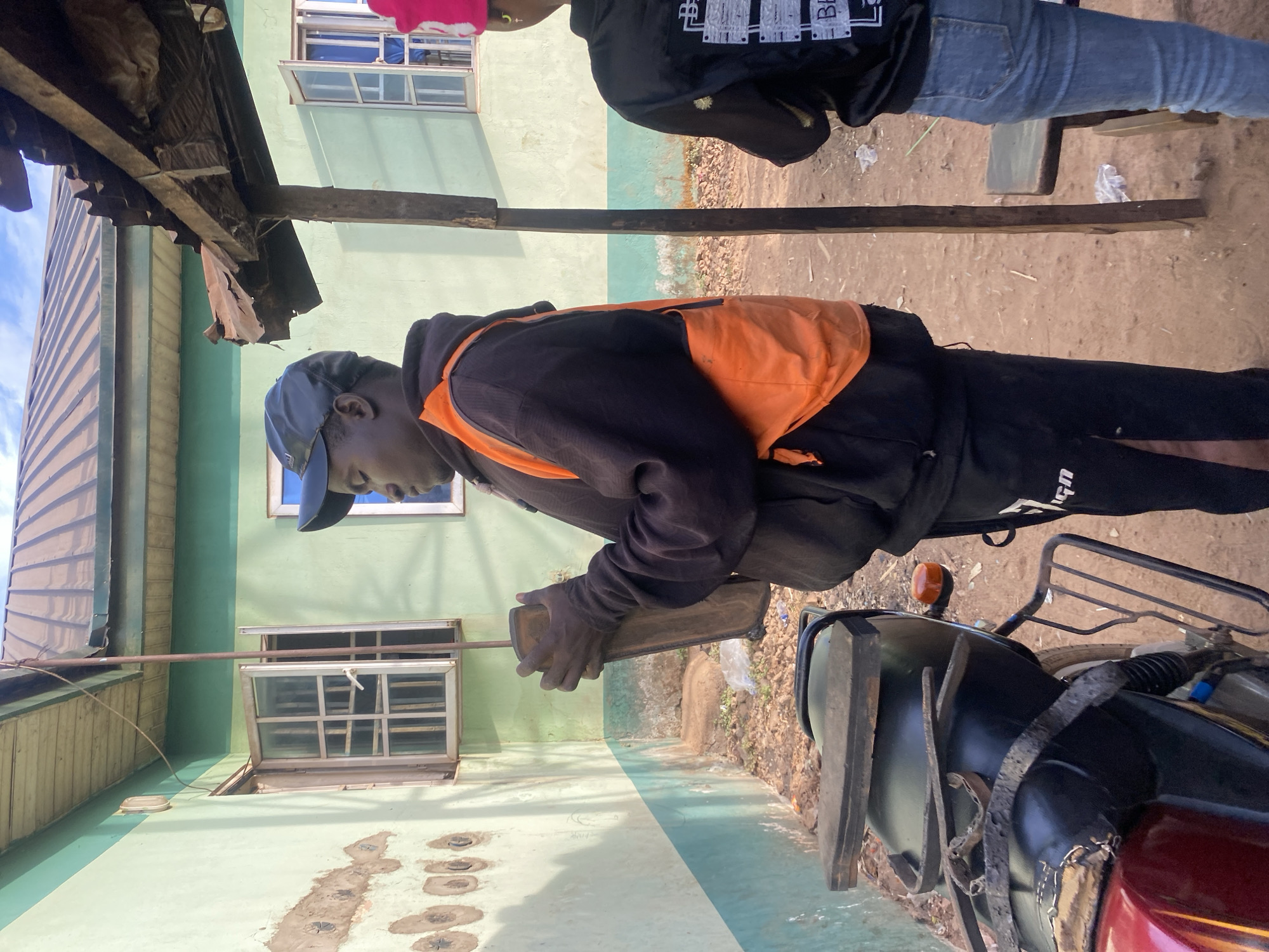 A man wearing a blue baseball cap, a black hooded sweatshirt with an orange vest, and black pants is positioned next to a black motorcycle. The man holds a rectangular object in his hands. The scene is set in an outdoor environment in Rido, Nigeria, in front of a light-green building with white-framed windows. Another person stands in the background, partially visible.