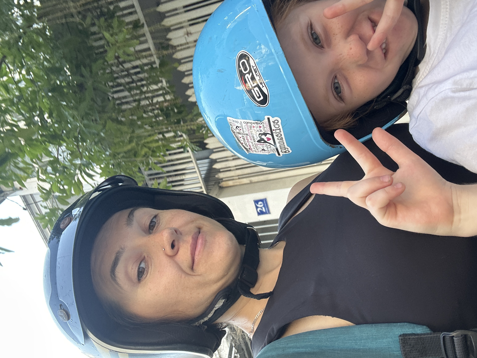 Two people are in a selfie on a scooter in front of a building with a numbered sign. The person in the foreground on the left is a woman wearing a black helmet and a black tank top. She is looking at the camera. The person on the right is a child wearing a blue helmet with stickers on it. The child is making a peace sign with their fingers. A green backpack is visible on the left side of the frame. The background shows a white fence and lush green trees. A blue sign with the number "26" is mounted on the wall of the building. The overall scene suggests an outdoor setting in a urban or suburban area.
