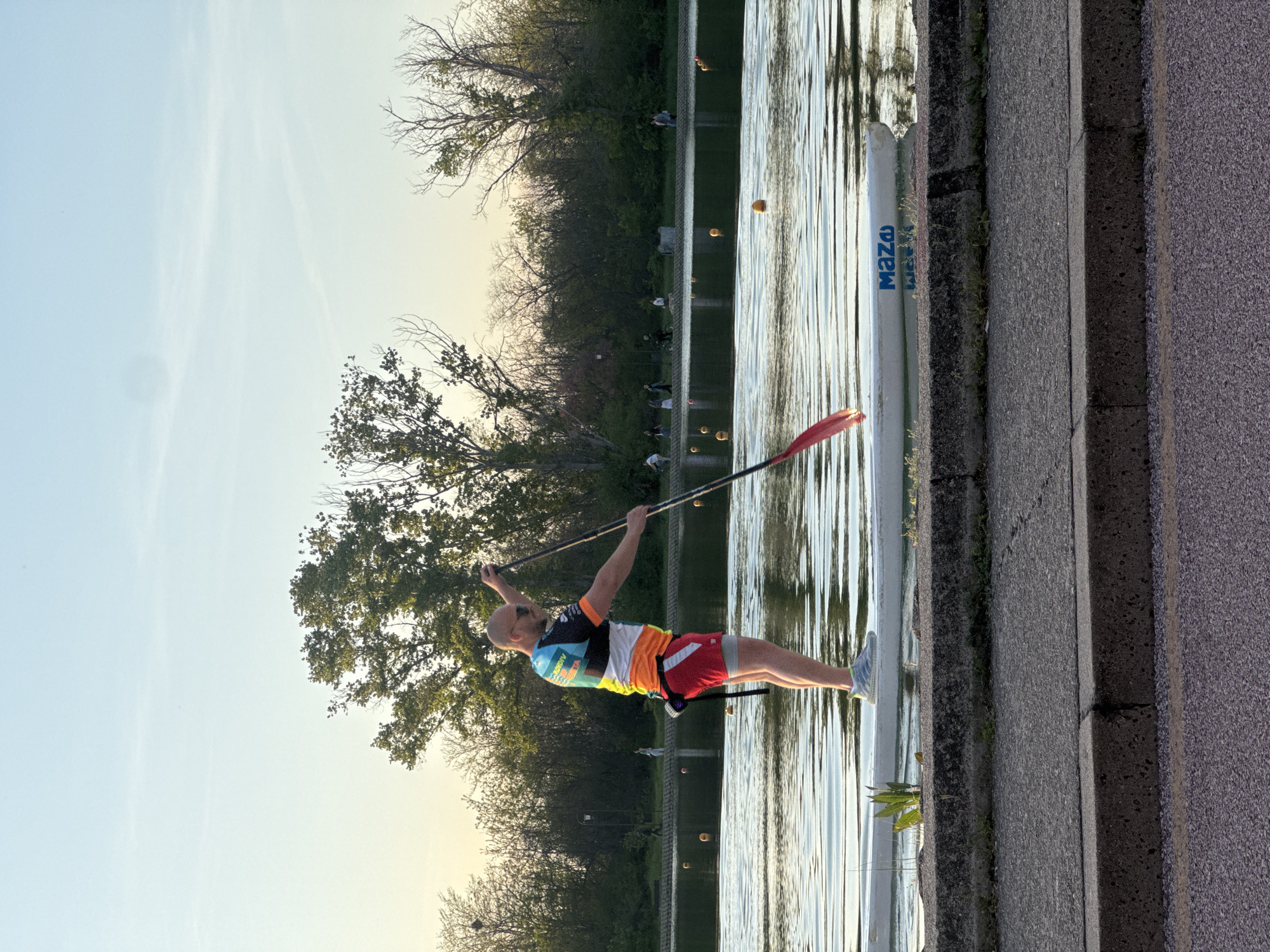 A male athlete is captured mid-action on a body of water, likely a canal or lake, in Plovdiv, Bulgaria. He stands upright on a narrow white watercraft, possibly a stand-up paddleboard or a competitive canoe/kayak, holding a long black paddle with a red blade. He is bald, has an athletic build, and wears a vibrant, multi-colored short-sleeved athletic top (featuring blue, yellow, orange, red, black, and white patterns) and red and orange shorts, paired with light-colored athletic shoes. His posture suggests focus and exertion, as if in the midst of a paddle stroke or preparing for one.
The water is calm with subtle ripples, reflecting the clear sky and distant trees. Several yellow buoys are visible floating in the water, suggesting a marked course or training area. The watercraft itself bears the visible text "MAZA" in blue lettering on its side. Along the edge of the water is a concrete bank or path. The background features a lush green tree-lined opposite bank, with some bare-branched trees also visible, and faint figures of people in the distance. The sky is a clear, bright blue with a slight gradient, indicating either early morning or late afternoon. The overall impression is one of active recreation or training in favorable weather conditions.