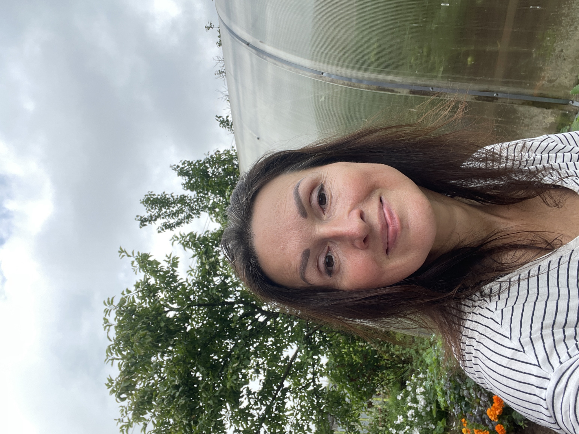 A selfie of a woman taken outdoors in Дресвяново, Russia. The woman, with brown hair and fair skin, is smiling. She is wearing a white shirt with black stripes. Behind her, to the left, is a green tree. To the right is a greenhouse. The sky is overcast.