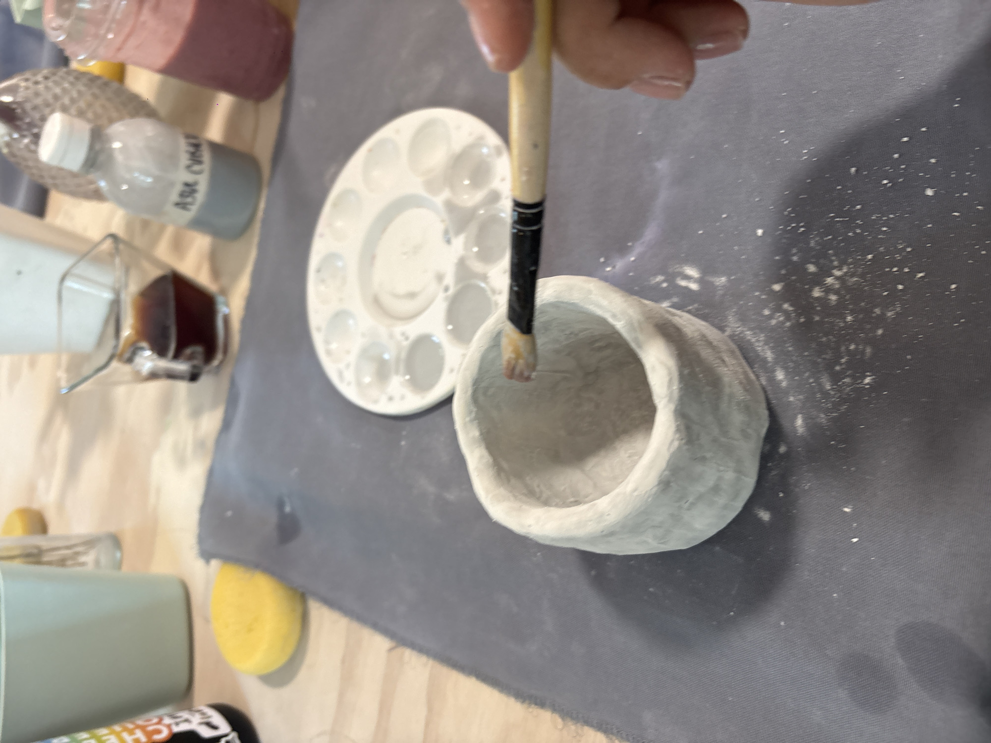 A close-up shot displays an artistic scene with a predominantly gray and white color palette. A hand holds a paintbrush over a small, white, textured bowl, as if in the process of painting. A white paint palette with numerous circular depressions is positioned to the left of the bowl.
Surrounding these central elements are various objects: bottles, a glass containing a dark liquid, a green cup, and a yellow sponge, all arranged on a surface covered with a gray cloth. The setting appears to be an indoor workspace, possibly an art studio or crafting area.
The image does not provide sufficient information to confirm the location or any specific details about the city of San Bernardino Tlaxcalancingo, Mexico.