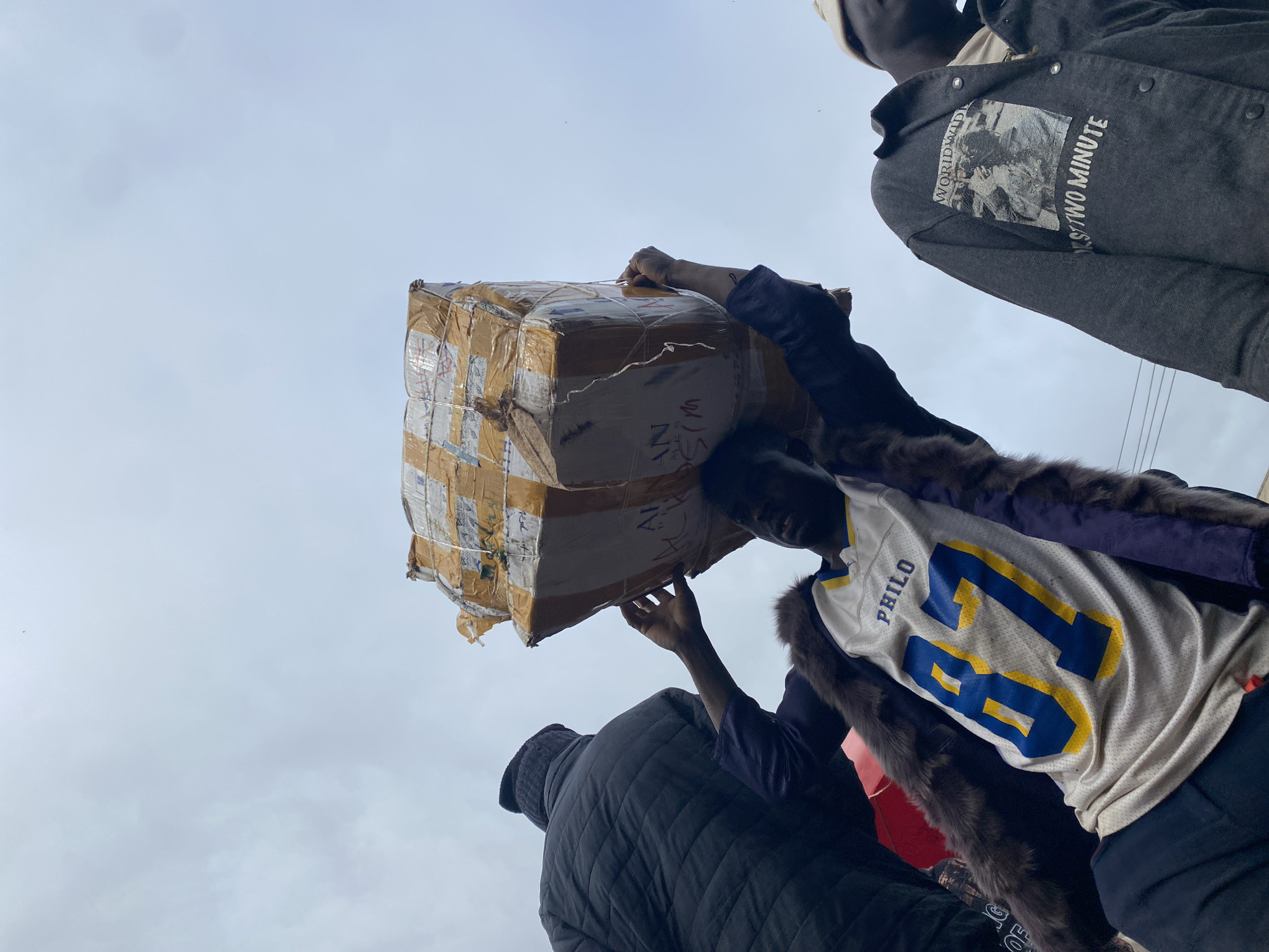 In Katon Rikkos, Nigeria, under an overcast sky, a young Black man, viewed from below, balances a large, taped cardboard box on his head. He wears a tan, sleeveless athletic jersey with the word "PHILO" and the number "87" in blue and yellow. A dark, fur-lined jacket is draped over his shoulders. To his left, another Black individual, also viewed from below, is partially visible, wearing a dark, quilted puffer jacket and a black knit hat. To his right, a third Black person wears a dark denim jacket with a graphic image and text that reads "WORLDWIDE" and "FOR STWO MINUTE." The scene appears to be outdoors, possibly in a busy area given the presence of multiple individuals and the apparent conveyance of goods.