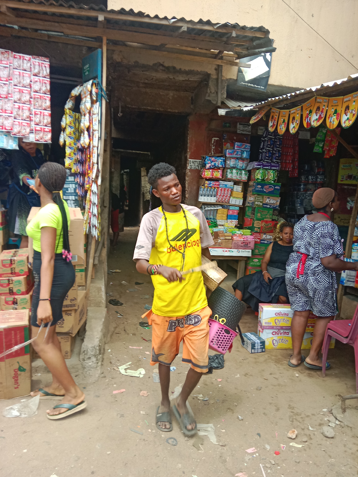 The image captures a street scene in Onitsha, Nigeria. At the center stands a young man wearing a yellow t-shirt with the word "Balenciaga", and orange shorts with "BEARD" printed on them. He holds a stick and what appears to be two baskets of different colors. To his left, a young woman with braided hair and a lime green top is walking away, a black purse slung over her shoulder, passing boxes of goods.
To the right of the young man is a small shop. A woman in a brown hat and patterned dress is standing near a red plastic chair. Another woman sits inside the shop, boxes of Chivita drinks piled nearby.
The shop is stocked with various items and displays promotional banners overhead. Other shop stalls are seen in the background. The ground is dirt or concrete, scattered with debris.