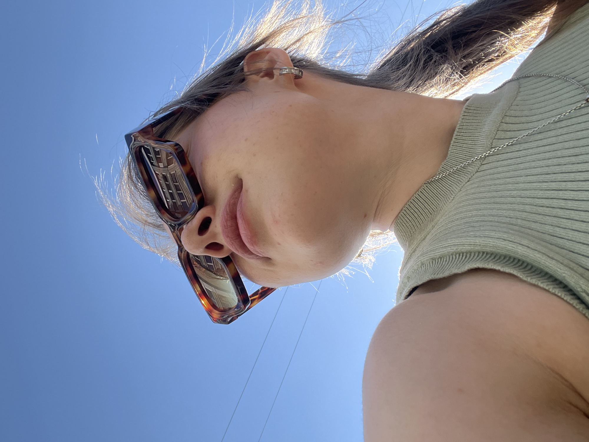 A low-angle, close-up photograph shows a person with sunglasses and an earring standing outdoors against a bright blue sky. The lenses of the person's sunglasses reflect a light-colored building with windows. The person is wearing a light-green ribbed shirt and a silver necklace. Utility wires extend across the sky in the background. This picture does not contain enough information to determine the location in the city of Chaikovsky, Russia.