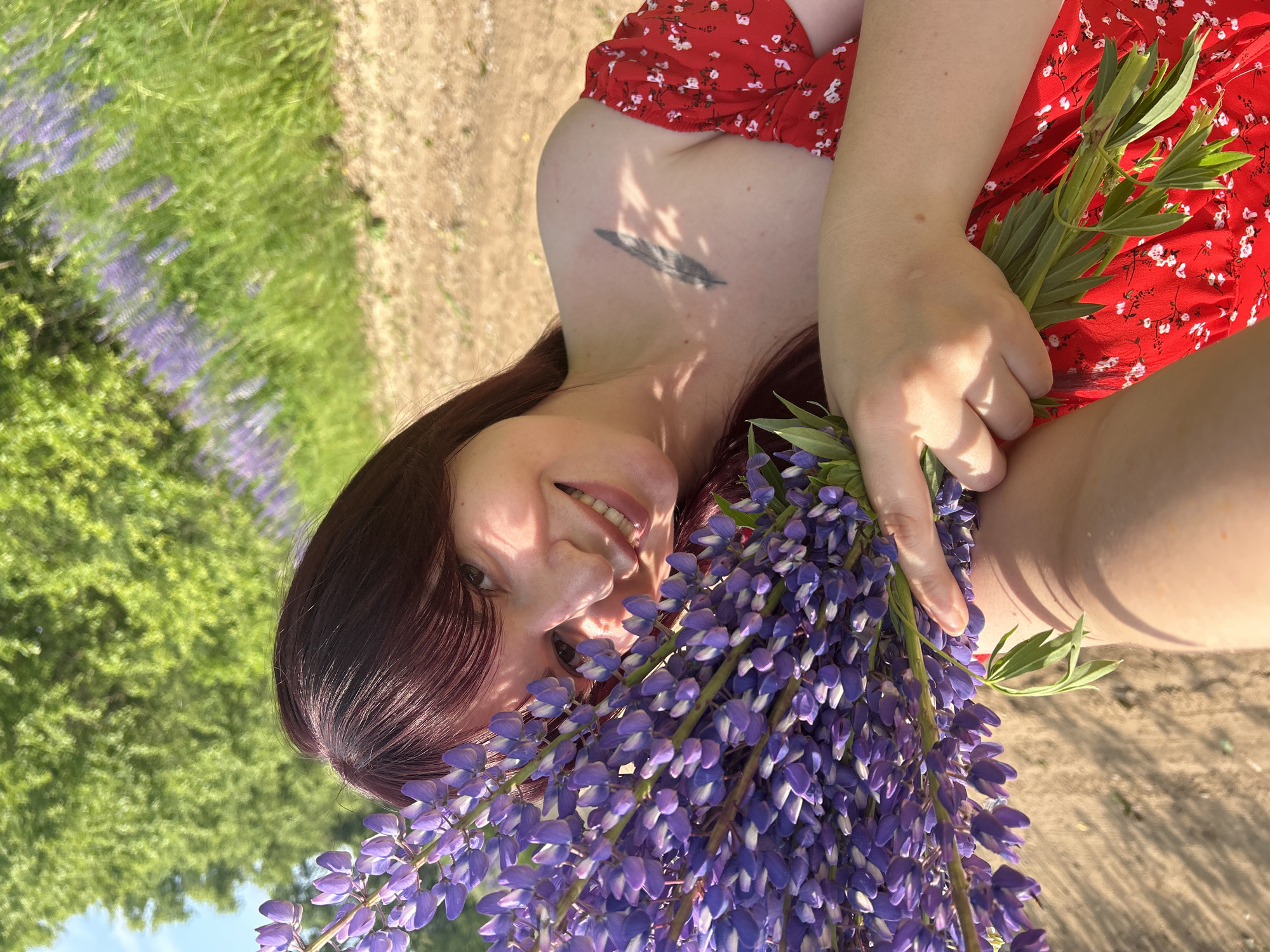 In an outdoor setting, a woman with dark red hair and a red floral dress is holding a bouquet of purple lupine flowers. She is smiling and looking towards the camera. A tattoo of a feather is visible on her left shoulder. The background consists of a sandy area, green trees, and a field of purple flowers. The scene appears to be in or near the city of Garevaya, Russia.