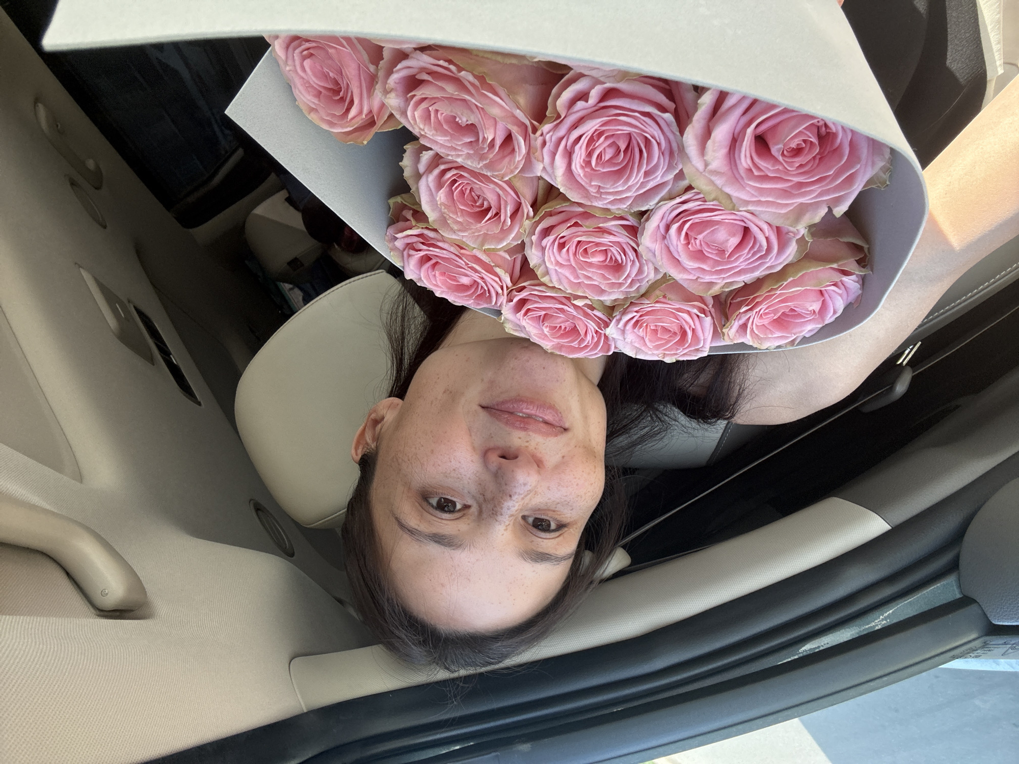 A woman with dark hair and freckles is holding a bouquet of pink roses in what appears to be the interior of a car. The car's interior is light-colored leather with a dark trim. The location appears to be in the city of Chaikovsky, Russia.