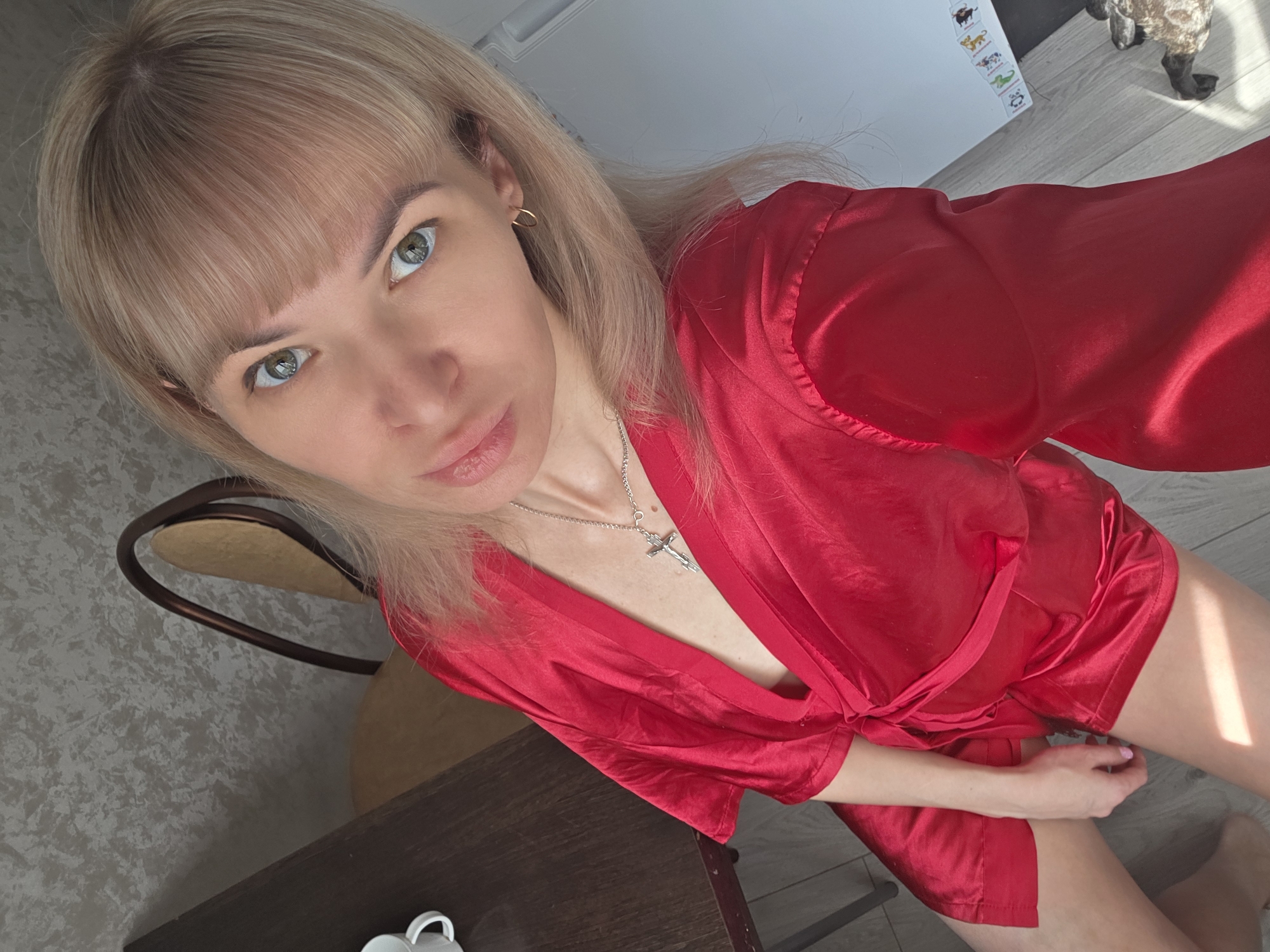 A young woman with blonde hair and bangs, and distinct greenish-blue eyes, is captured in a close-up, high-angle selfie. She has fair skin and is wearing a vibrant red satin robe, tied loosely at the waist. A silver necklace with a crucifix pendant adorns her neck, and a small gold hoop earring is visible in her left ear. She is looking directly at the camera with a neutral, composed expression.
The setting appears to be an indoor space, likely a home in Chaikovsky, Russia. The woman is positioned on a light wooden or laminate floor, next to a dark wooden table where a white mug is partially visible. Behind her, a chair with a rounded, dark frame is seen against a textured, light grey wallpapered wall. Bright natural light, suggesting daytime and sunny weather, illuminates the scene and casts soft shadows. In the upper right background, on the floor, a figurine resembling a sheep or similar animal can be seen. Behind her to the left, a white cabinet or refrigerator is adorned with several colorful animal stickers, including a bull, lion, and alligator. Below these stickers, two recycling symbols are visible, one containing the number "4".