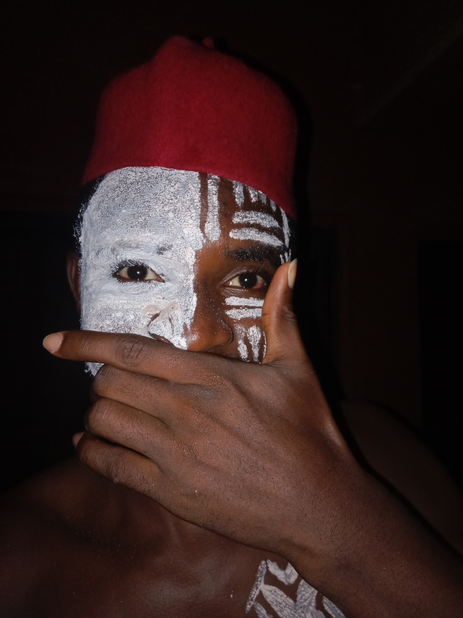 A close-up photograph captures a person in Unknown, Unknown. The person's face is partially covered in white paint, with distinct patterns extending onto the forehead. They are wearing a red hat. Their hand is raised to their mouth, partially obscuring it. The background is dark, suggesting a dimly lit environment.