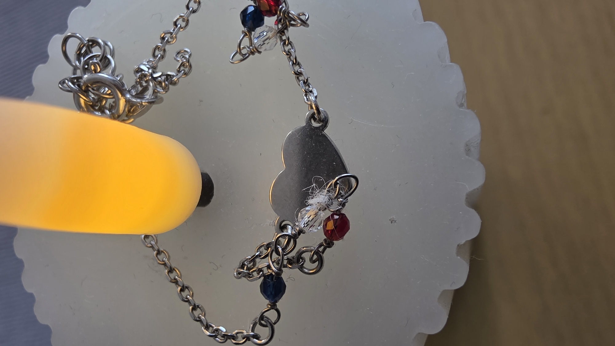 The image shows a close-up of a silver-colored charm bracelet with a heart pendant, clear and colored beads. The bracelet is resting on a white, scalloped surface. A bright, yellow light source illuminates part of the bracelet and the surface. The background is a light brown color. There is no information to suggest the photo was taken in Blagoevgrad, Bulgaria.