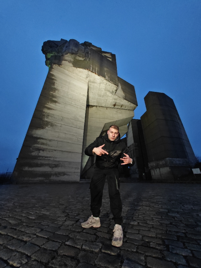 A single male subject is depicted standing on a cobblestone surface at the Monument to 1300 Years of Bulgaria in Shumen, Bulgaria, during what appears to be late evening or night. The sky is a deep, uniform blue, indicative of twilight or night conditions.
The male, in the foreground and center, faces directly towards the camera. He is wearing a dark, possibly black, hooded jacket with a subtle pattern on the chest, dark cargo-style pants, and light-colored athletic shoes with dark soles. His right arm is bent at the elbow, with his index and middle fingers extended and pointing downwards, while his thumb is also extended. His left arm is also bent at the elbow, with his index and pinky fingers extended upwards, forming a "rock on" or "horns" gesture, while his middle and ring fingers are curled. His expression is serious.
Behind the subject, the massive, brutalist concrete structure of the Monument to 1300 Years of Bulgaria dominates the background. The monument features angular, monolithic concrete slabs and towering elements. Portions of the monument are illuminated by artificial light, casting glows on its surfaces, with some sections appearing darker due to shadows. A faint green light is visible on the upper left section of the monument. The ground beneath the subject and extending into the foreground consists of dark, uneven cobblestones.