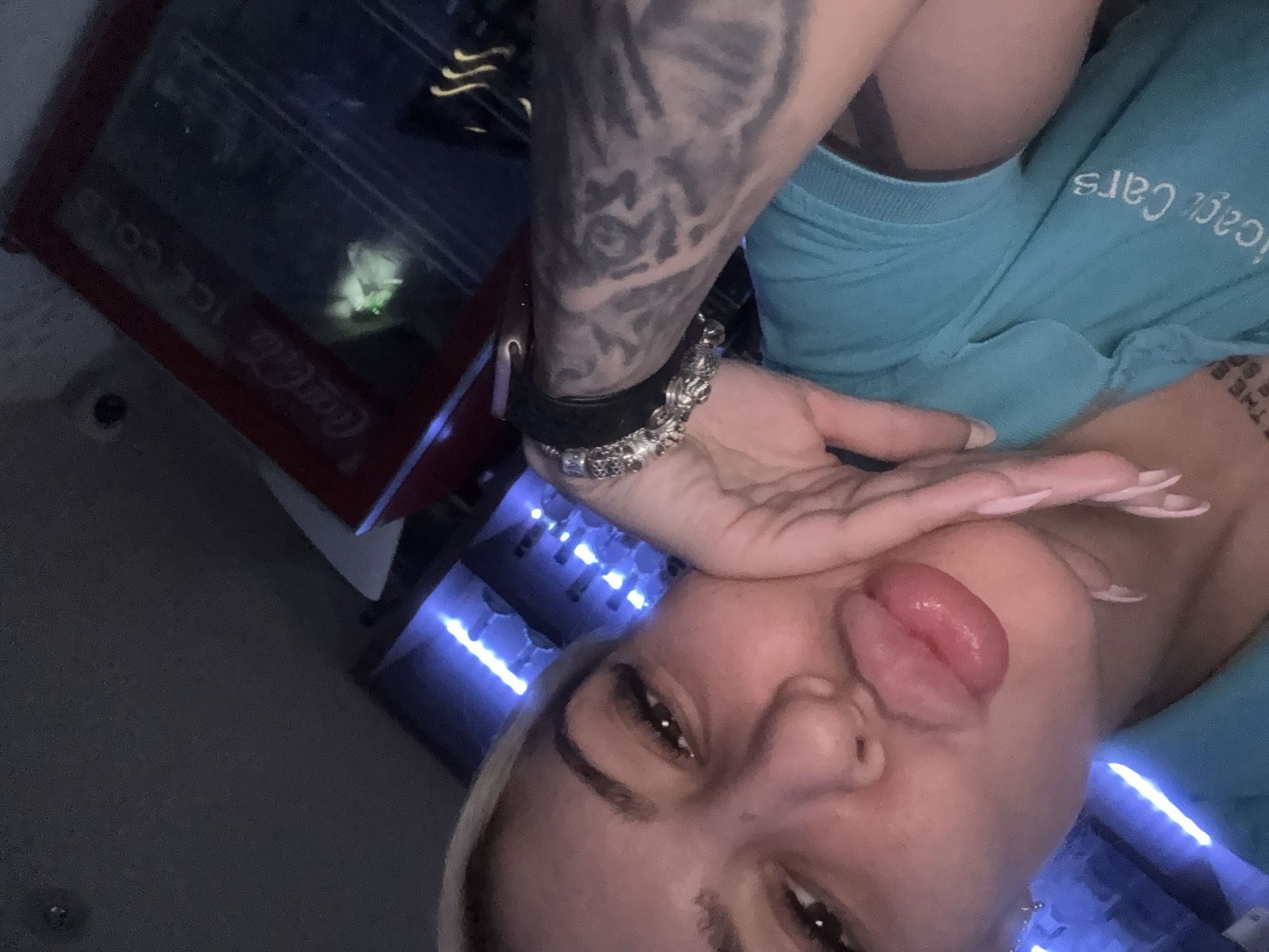 A person with light complexion, blonde eyebrows, and dark eyes is posing for a selfie. Their lips are pursed. Their right arm, heavily tattooed, is raised. A bracelet is visible on the wrist. The person is wearing a light blue top with white lettering. The background includes a Coca-Cola branded refrigerator, a keyboard with blue LED lights, and other equipment. The location cannot be identified as Plovdiv, Bulgaria.