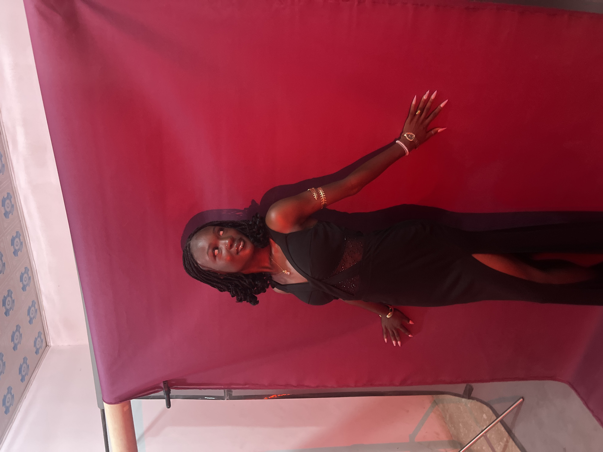 A young Black woman with dark braided hair stands in front of a magenta backdrop, with a red light illuminating the scene from the left. She is wearing a black, floor-length gown with a V-neckline and a thigh-high slit. The dress features a sheer, beaded panel across the midsection. She adorns herself with gold arm bands on both upper arms, a gold necklace, and a wristwatch on her left wrist, accompanied by a ring. Her fingernails are painted a vibrant pink. Her gaze is directed to her right, with a slight smile. The backdrop is draped on a stand, and the room appears to be a studio setting, possibly in Gura Top, Nigeria. A light stand is visible to the left of the frame, partially obscured by the backdrop.