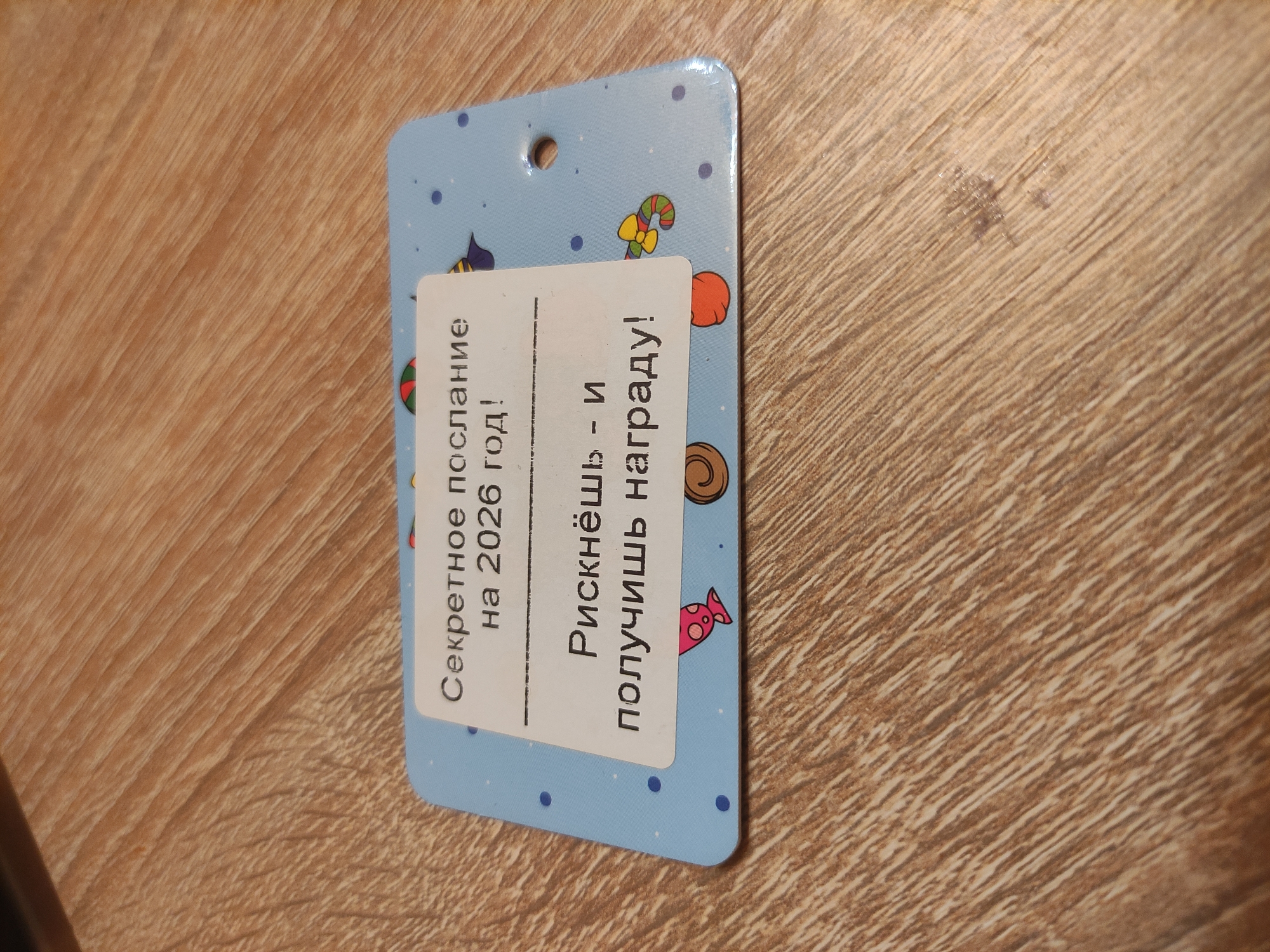 A light blue, rectangular, laminated card with rounded corners lies at a slight angle on a light brown wooden surface, exhibiting a distinct wood grain pattern. The card features a white rectangular label centrally placed, displaying Russian text in black lettering. The upper line of text reads "Секретное послание на 2026 год!" (Secret message for 2026!). Below a horizontal dashed line, the text continues "Рискнёшь - и получишь награду!" (Take a risk - and you will receive a reward!). The card's blue surface is decorated with scattered darker blue polka dots and small, cartoon-style illustrations, including a red fish, a striped candy cane, a brown coiled shape, and an orange mushroom-like figure. A single circular hole is punched through the card near the top right corner. The scene is devoid of people or direct action. No information within the image specifies a location in the city of Kratovo, Russia.