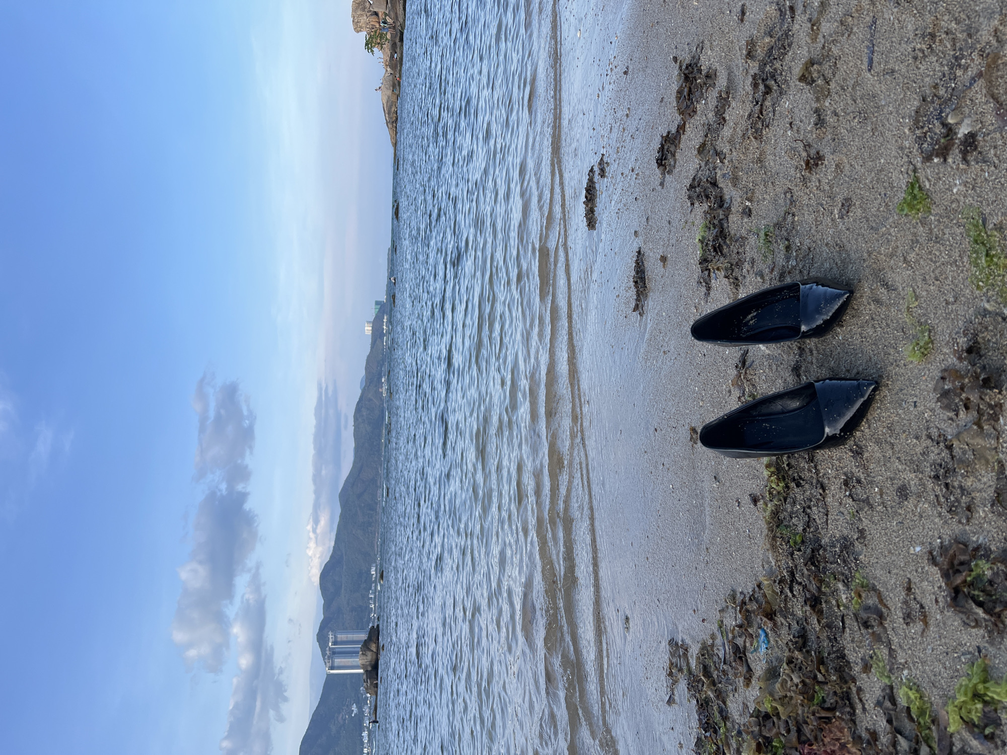 This image captures a serene coastal scene in Bac Nha Trang Ward, Vietnam, presented from a low angle looking out over the sea and shoreline. The composition is split between the sky, the sea, and the sandy beach.
In the foreground, two black flippers or water shoes are placed on the damp sand at the water's edge. The sand itself is a mix of wet and dry areas, with visible ripple marks left by receding waves. Patches of green and brown seaweed, along with small stones and debris, are scattered along the tideline, indicating a natural, somewhat rugged beach.
The calm sea occupies a significant portion of the mid-ground, displaying gentle ripples across its surface, reflecting the bright sky above. In the distance, along the horizon, a line of mountains or hills frames the coastline. Several modern high-rise buildings, possibly hotels or residential complexes, are visible nestled against the mountains and along the water, suggesting urban development near this natural setting.
The sky above is predominantly clear and bright blue, with some scattered white cumulus clouds, particularly towards the left of the frame. The overall lighting suggests it is daytime, likely late morning or early afternoon, under fair weather conditions. No specific human activity is depicted, and no identifiable people are clearly visible, contributing to a tranquil and undisturbed atmosphere. There is no visible text within the image.