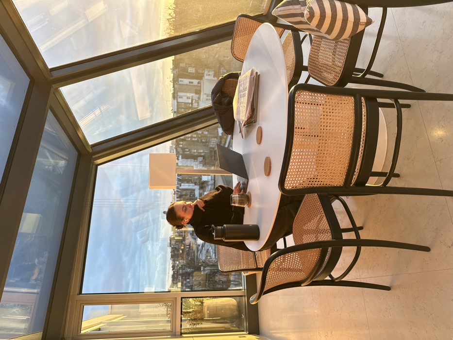 A person sits at a round, white table surrounded by rattan chairs in a sunlit room with large windows. Outside, buildings and a sky transitioning from day to dusk are visible. On the table, a laptop, a water bottle, a glass of liquid, coasters, and a newspaper are placed. The person, wearing a dark garment, is engrossed in their work. The room is illuminated by natural light pouring in through the angled windows.