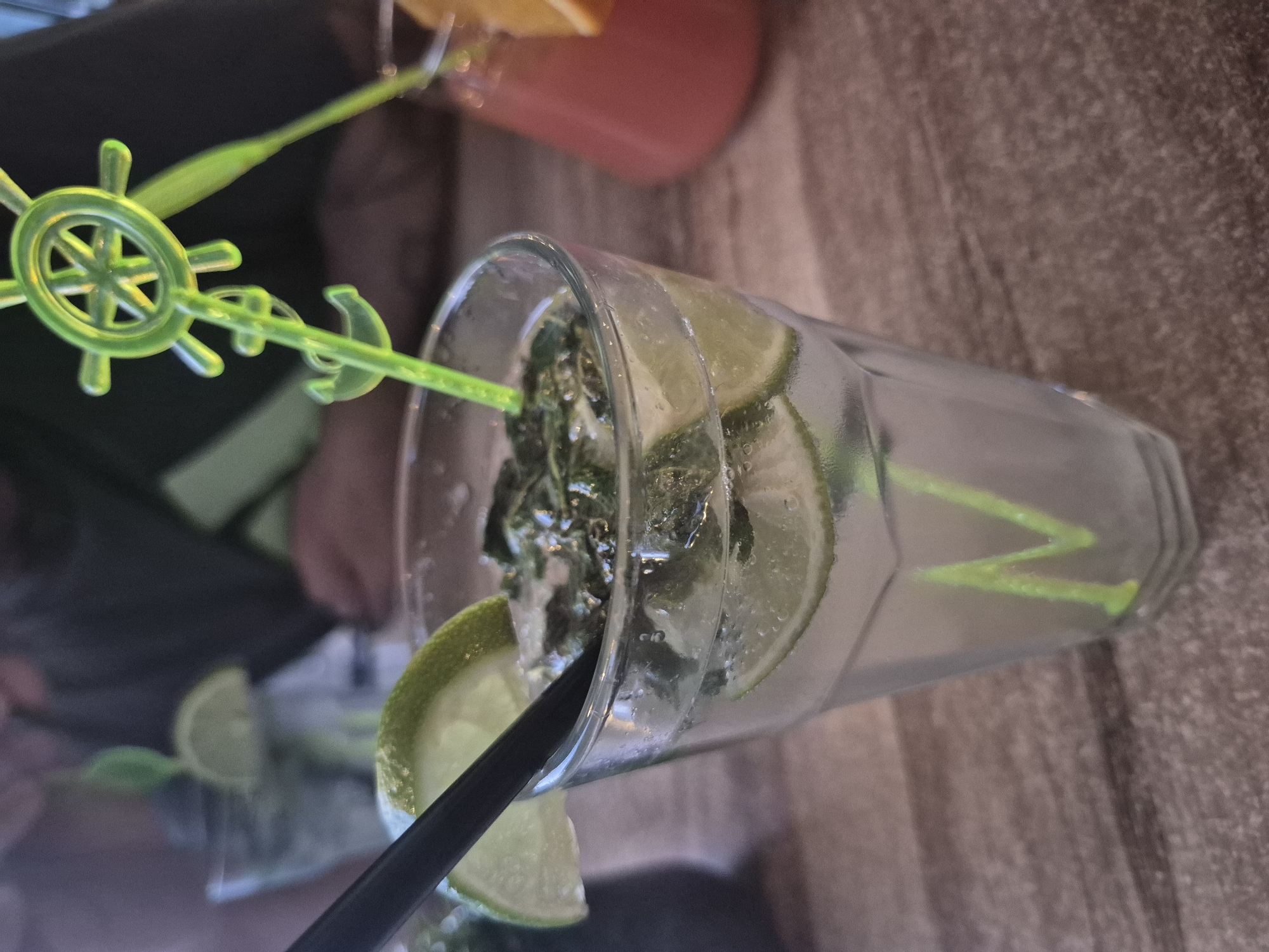 A close-up, canted shot shows a cocktail with lime wedges and a straw in a glass. The drink is on a surface with a textured, slightly blurred background. A second drink in a pinkish hue is visible in the background with a green plastic decoration of a ship's wheel and anchor. There is no visible evidence to indicate a specific location in Pavlikeni, Bulgaria.