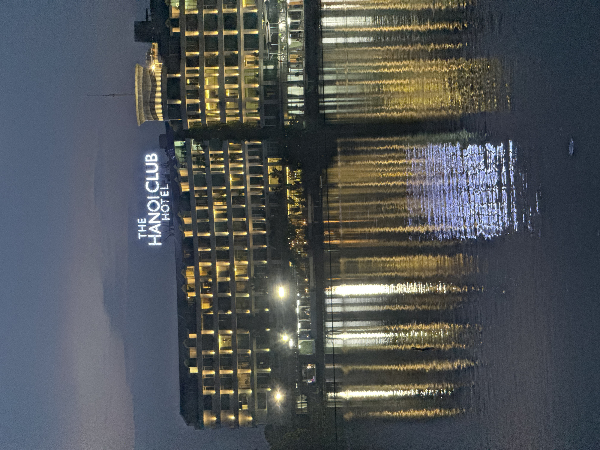 The image shows The Hanoi Club Hotel in Hanoi, Vietnam, at dusk. The hotel is a large, multi-story building with many windows illuminated by warm yellow light, reflecting in the calm water in front of it. A large, glowing sign on the roof reads "THE HANOI CLUB HOTEL." The sky is a dusky blue with some clouds. The water reflects the lights from the hotel, creating a shimmering effect. The scene is serene and peaceful, with no people clearly visible. The architecture of the building suggests a modern design, with clean lines and a symmetrical facade. The overall impression is one of elegance and tranquility, capturing a beautiful moment in the city.