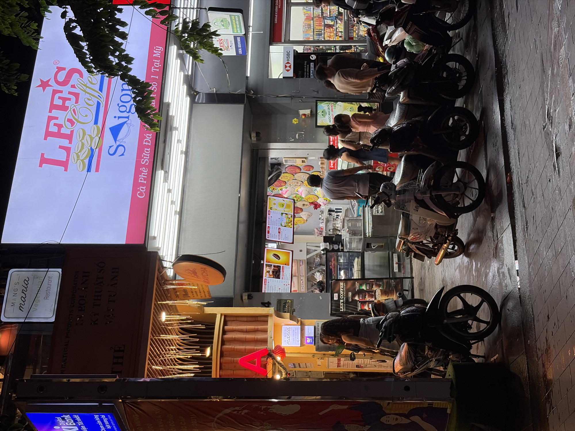 The image shows a street scene in Thủ Đức, Vietnam, at night. Several motorbikes are parked along the sidewalk in front of a row of businesses. "Lee's Coffee" has a large, illuminated sign with the Vietnamese phrase "Cà Phê Sữa Đá". Next door is a "MANG'S mania Vegetarian Restaurant" with a lighted sign and decorative wheat stalk design. People are visible inside Lee's Coffee, some appearing to order drinks. ATM is visible next to a HSBC sign. Across the street, a partial advertisement is visible.