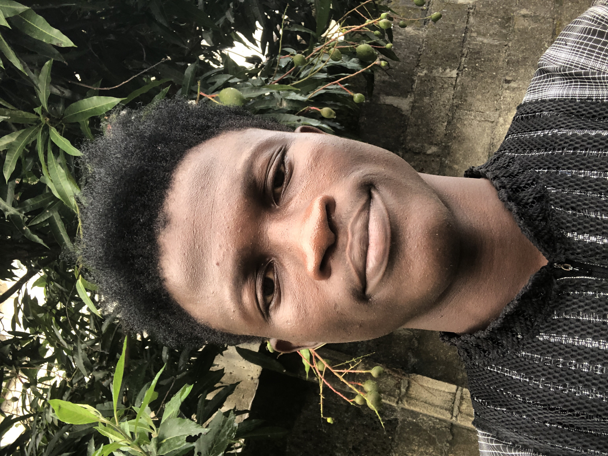 A young Black male is pictured from the chest up, posing for a selfie outdoors. He has dark skin, short, black, curly hair, and is looking directly at the camera with a neutral expression. He is wearing a black shirt with a textured pattern and a zipper. The background consists of lush green foliage and a concrete wall, suggesting a tropical environment. The overall impression is a casual portrait taken in a natural setting, potentially in Ajah, Nigeria, given the hint of a mango tree with developing fruit.