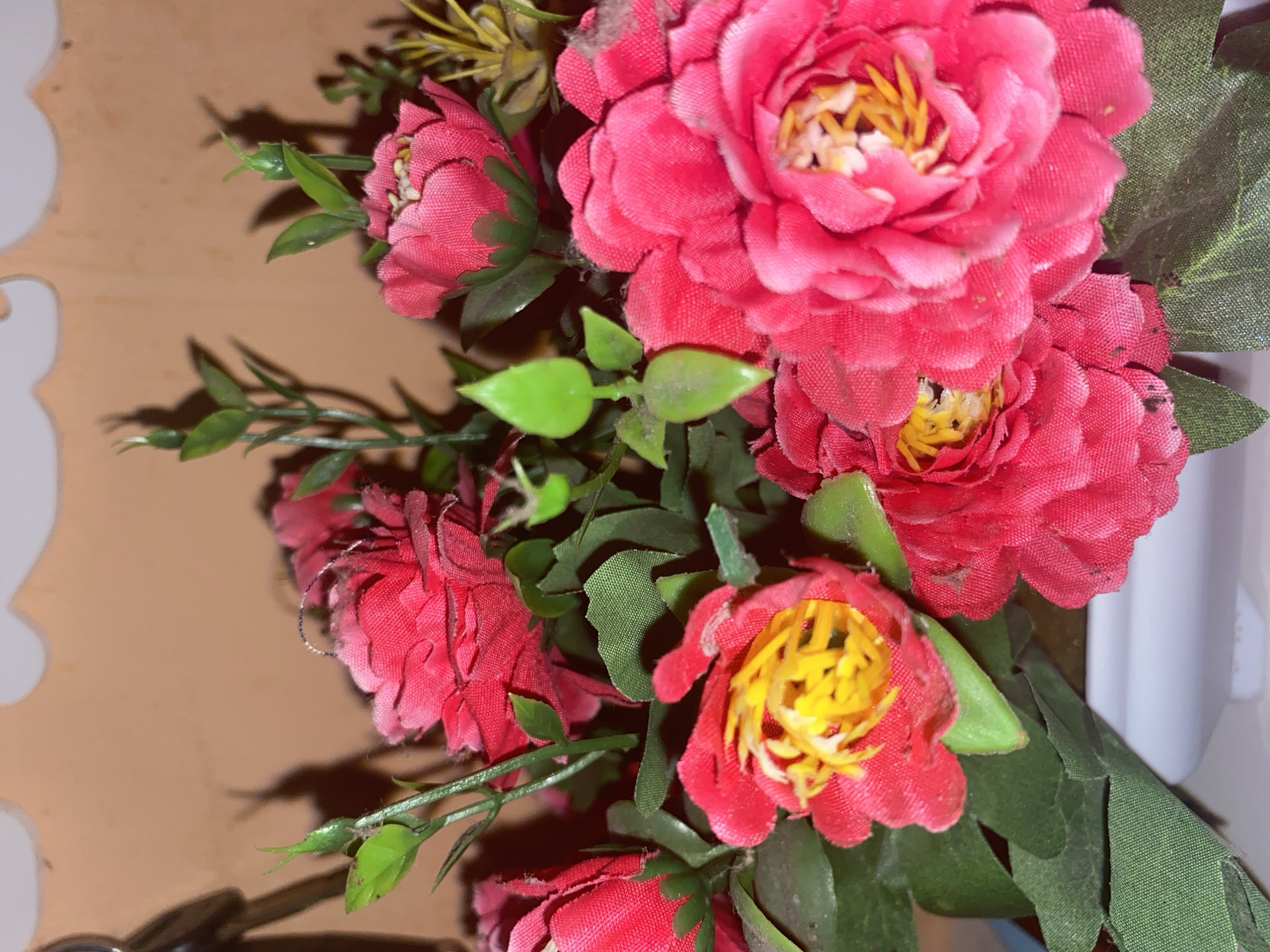 A close-up, rotated shot shows a bouquet of artificial pink flowers with yellow centers. The flowers are clustered together, with green leaves and stems interspersed. The background is a muted brown, with some lighter, irregular shapes visible on the left side of the frame, possibly part of a decorative item or furniture. Shadows are cast by the flowers and leaves onto the background, suggesting a light source from the top right. The overall lighting is warm. There are no people, text, or discernible weather conditions. The image focuses solely on the floral arrangement.