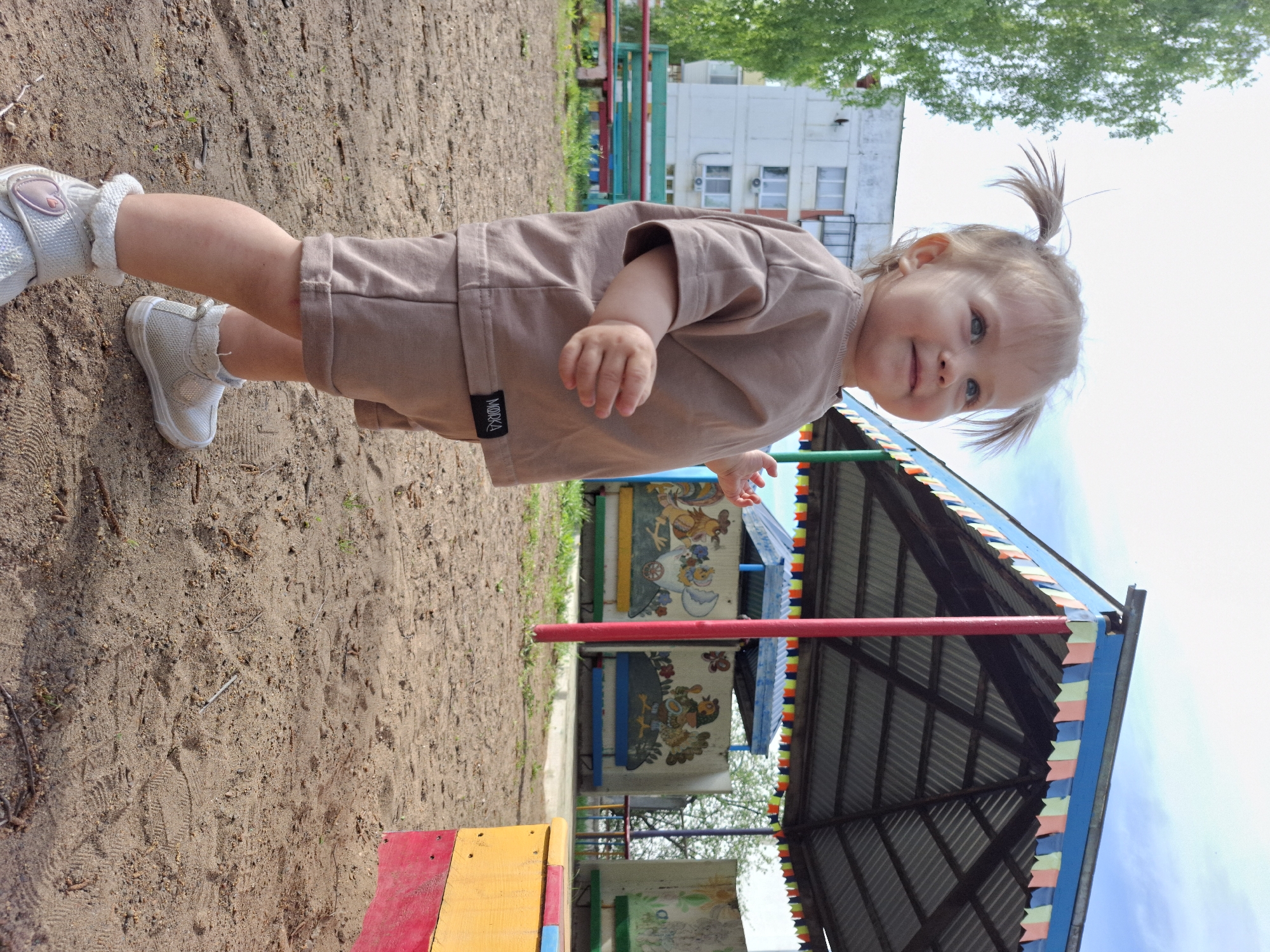 A full, slightly rotated shot shows a young child at a playground in Chaikovsky, Russia. The child, wearing a light brown shirt and shorts set and white shoes with ankle detailing, stands on a sand-covered ground. The child has light hair pulled into two small ponytails and is looking towards the camera. To the right and behind the child is a structure with a corrugated metal roof, painted red poles, and colorful painted panels on the walls. In the background, there is a multi-story building with windows, visible foliage, and green playground equipment. The sky is visible at the top right of the image.