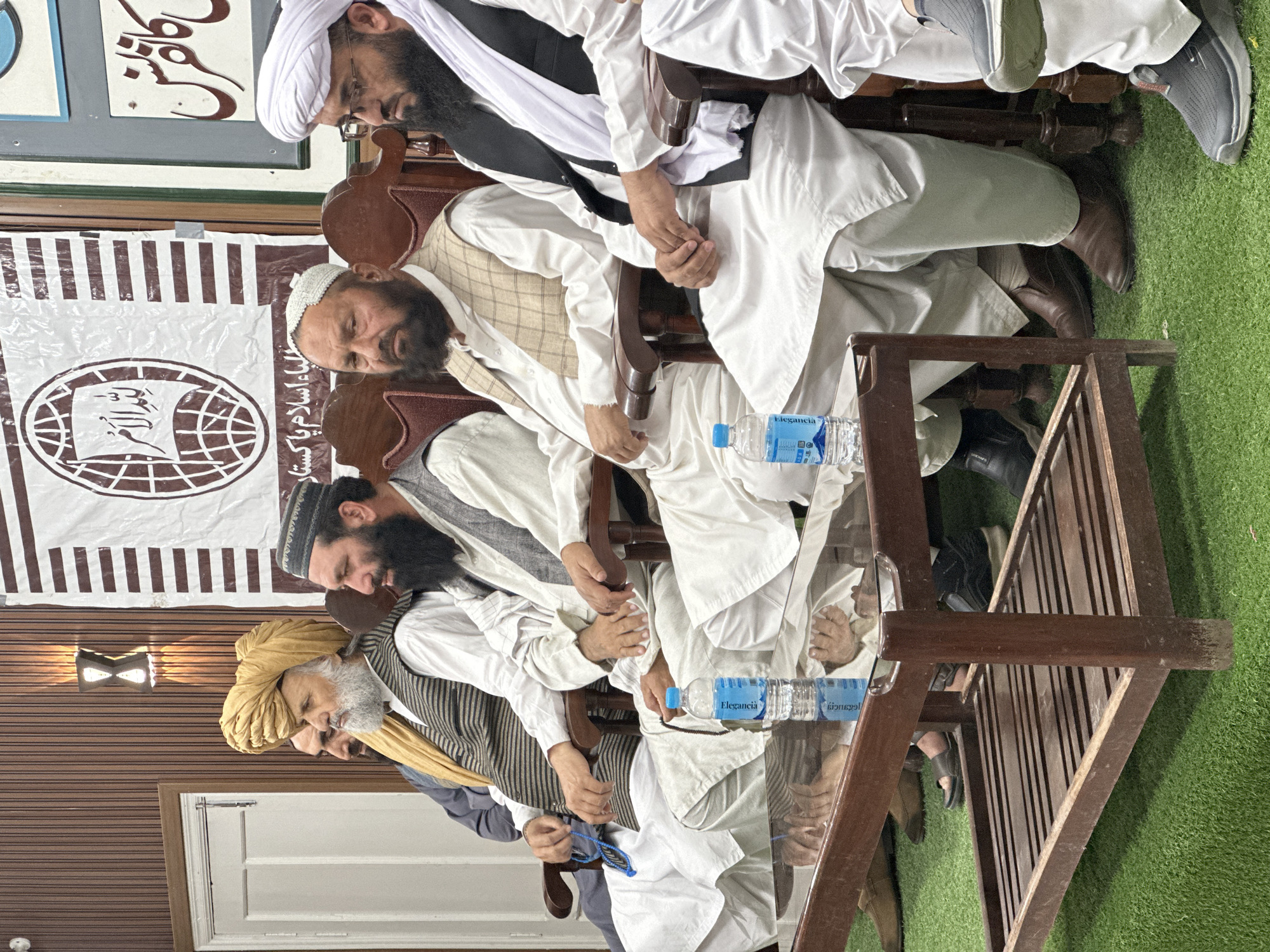 Four men are seated next to each other at a table in an indoor setting, with a banner visible in the background. The men appear to be of Pakistani or Afghani descent and are likely Muslim clerics or religious leaders. They are all wearing traditional attire, including turbans, vests, and long, loose-fitting robes. Their beards are long and well-groomed.
The table in front of them holds two bottles of "Elegancia" brand water. The room has a wooden interior, including walls with horizontal stripes and ornate wall sconces. A white door is visible on the left. A green carpet mimicking grass texture lies on the floor. Based on the banner and contextual clues, the location is likely in Quetta City Tehsil, Pakistan, possibly a religious institution or meeting place. The men appear to be engaged in a formal gathering or discussion.