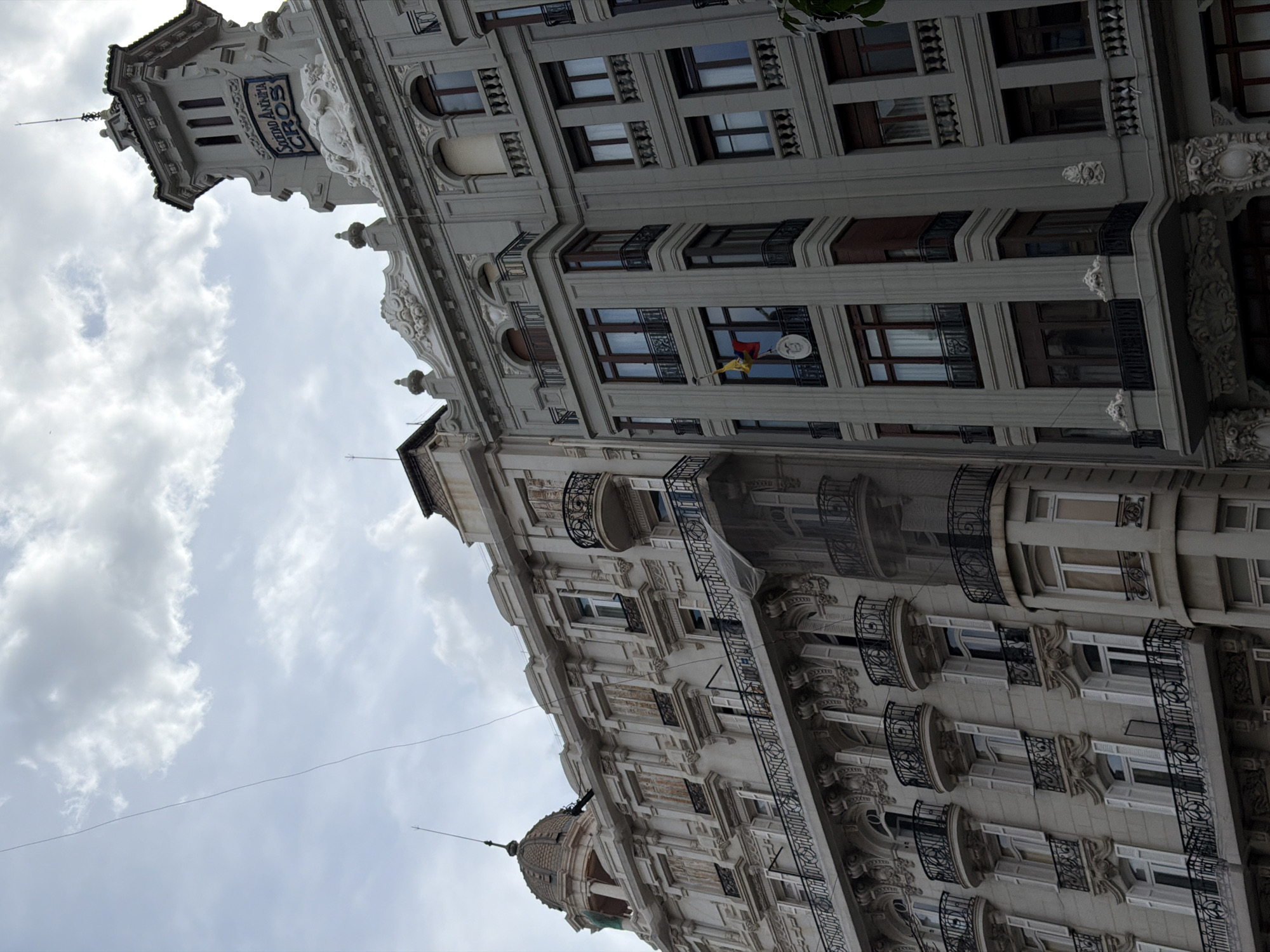 The image displays an upward perspective of two historically significant, ornate buildings located on Carrer de Colón in Valencia, Spain. The sky is overcast with scattered clouds.
The building on the right is a multi-story structure with a grey facade, characterized by classical architectural details. Its prominent feature is a square tower at the upper right, adorned with a blue sign bearing the white text "SOCIEDAD ANÓNIMA CROS." Below the tower, the building exhibits decorative cornices and balustrades. Multiple windows, framed in dark wood, are visible across its floors, some accompanied by small wrought-iron balconies. From a balcony on an intermediate floor, a vertically suspended flag of Ecuador is visible, featuring wider yellow, blue, and red stripes with a central coat of arms.
To the left, a multi-story building with a lighter, beige-colored facade stands adjacent. This structure is highly decorative, featuring numerous windows with white louvered shutters, many of which are closed. Intricate sculptural elements adorn the window surrounds and balustrades. Ornate wrought-iron balconies extend from several windows. A distinctive dome structure is visible on the building's roof towards the upper left. A translucent mesh net is draped over a section of the facade of this lighter-colored building.
A single thin, dark wire traverses the sky diagonally from the left side of the frame towards the upper right. No individuals are present in the scene. The architecture of both buildings suggests an early 20th-century construction period, combining eclectic and classical elements.