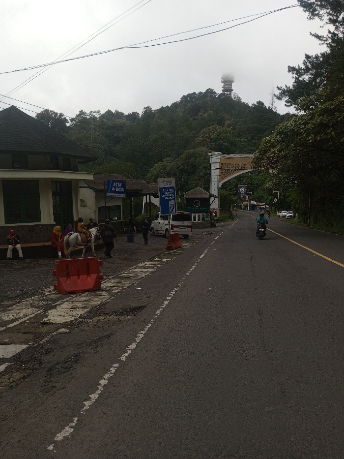 A road leads towards a mountainous area, featuring an archway structure. A communications tower is visible atop the hill, partially obscured by clouds. A building sits on the left, near parked vehicles and people. A horse stands near orange barriers. A motorbike travels along the right side of the road, alongside a yellow line. ATM and Electric Vehicle Charging Station signs are also present.