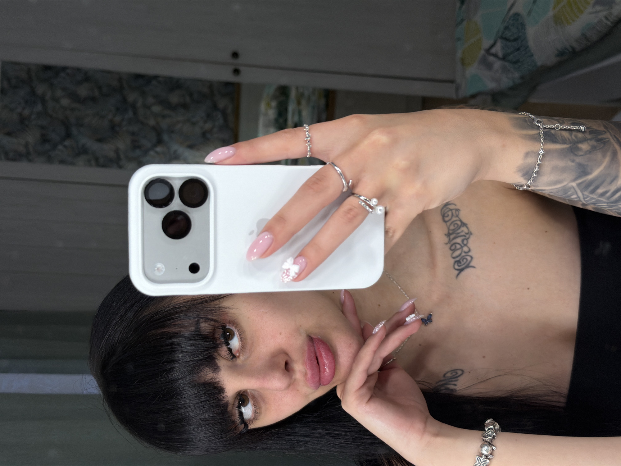 A young woman with dark, straight hair and bangs is depicted taking a selfie in a mirror. She has dark eyebrows, long eyelashes, light brown eyes, and full, glossy pink lips. Her left hand, adorned with several silver-colored rings and light-colored nails (some with glitter or French tips), rests under her chin. Her right hand holds a white smartphone with a three-lens camera module and flash in a white case, positioned to capture the image. This hand also features light pink, almond-shaped nails, with a white 3D flower embellishment on the ring finger, and several silver-colored rings. She is wearing a black strapless top. Visible tattoos include dark script or ornamental designs on her upper chest/collarbone area and a shaded sleeve tattoo on her right forearm. A silver charm bracelet is on her left wrist, and a silver-colored chain bracelet with decorative elements is on her right forearm. A silver-colored necklace with a dark blue butterfly pendant is around her neck. The background, largely out of focus, shows a reflective surface consistent with a mirror, revealing hints of interior architecture like a door or wall and a patterned textile with green, yellow, and blue tones.