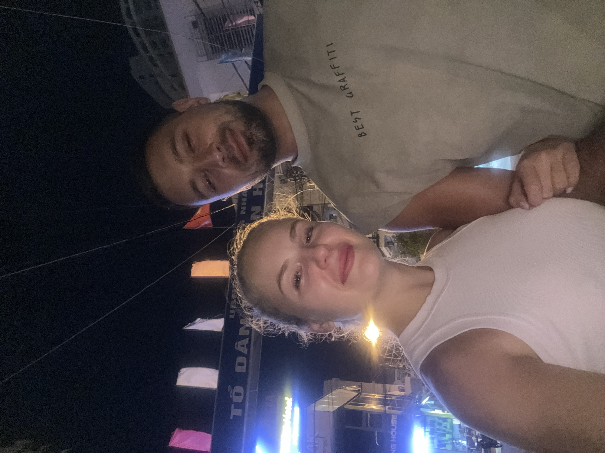 A low-angle, full-shot photograph captures a white couple posing for a selfie at night in Nha Trang, Vietnam. The woman, on the left, is fair-skinned with blonde hair styled in a messy bun. She wears a white sleeveless tank top. Her eyes are closed, and she has a slight smile. The man, on the right, has tanned skin and short dark hair with stubble. He wears a light olive-green sleeveless t-shirt with the words "BEST GRAFFITI" printed on it in black. He looks at the camera with a smile. Behind them, a street is visible with illuminated signs, including "TỔ DÂN..." and "NG HOUSE," indicating their location is likely a commercial area. Several colorful flags are strung across the street, and a building with multiple floors can be seen in the background. The lighting is artificial, creating a nighttime urban ambiance.