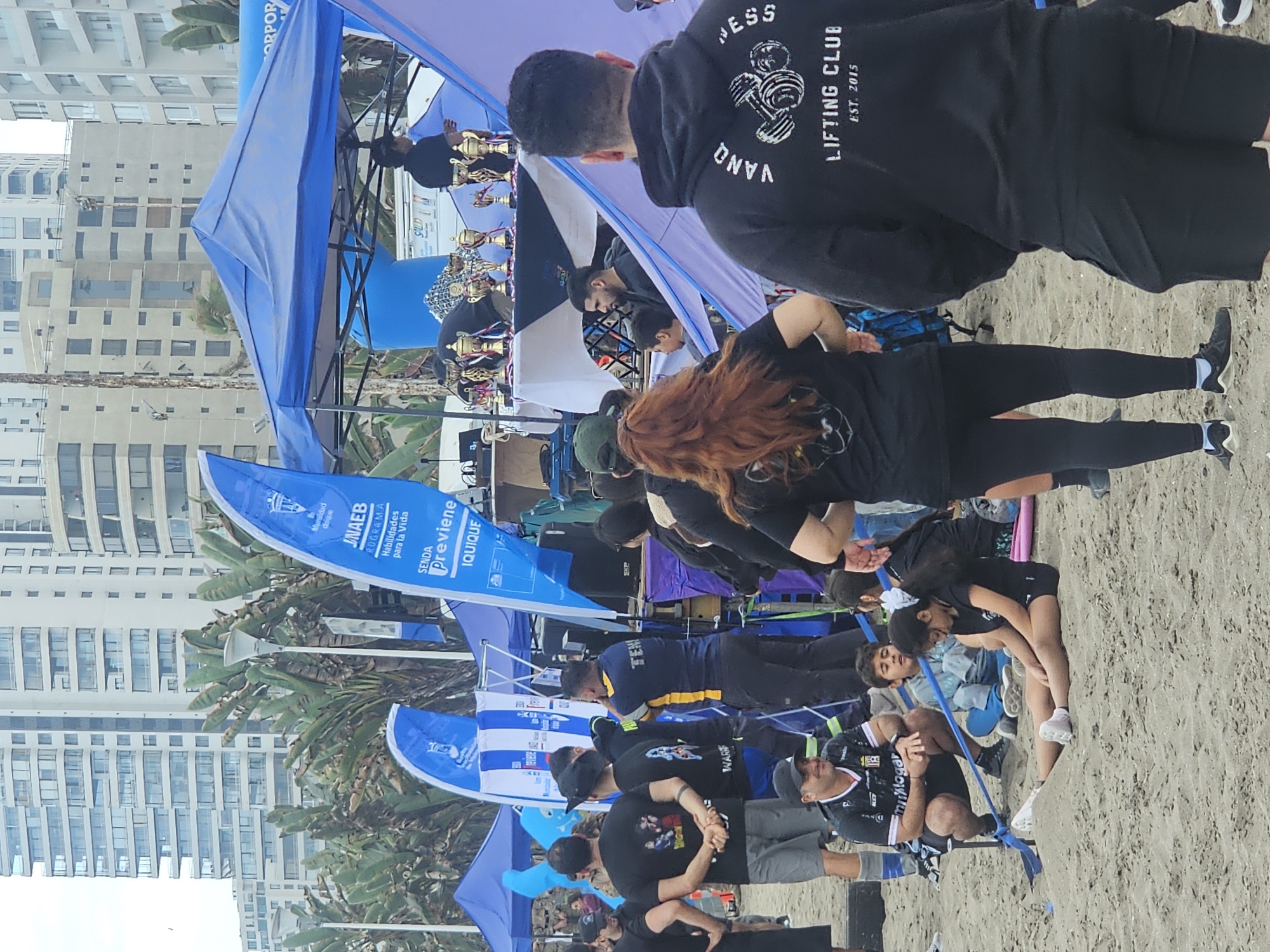 The image depicts an outdoor event on a sandy beach, likely in Iquique, Chile, given the presence of flags bearing "IQUIQUE" and "SENDA Previene." A crowd of people, diverse in age and gender, are gathered around tents and a display of trophies. Several individuals are wearing athletic attire, and some have logos or text on their clothing, suggesting a sporting or community event. In the background, tall, modern buildings are visible, indicating an urban coastal setting. The environment includes sandy ground, palm trees, and a cloudy sky. There is a sense of organized activity, with people standing, sitting, and observing.