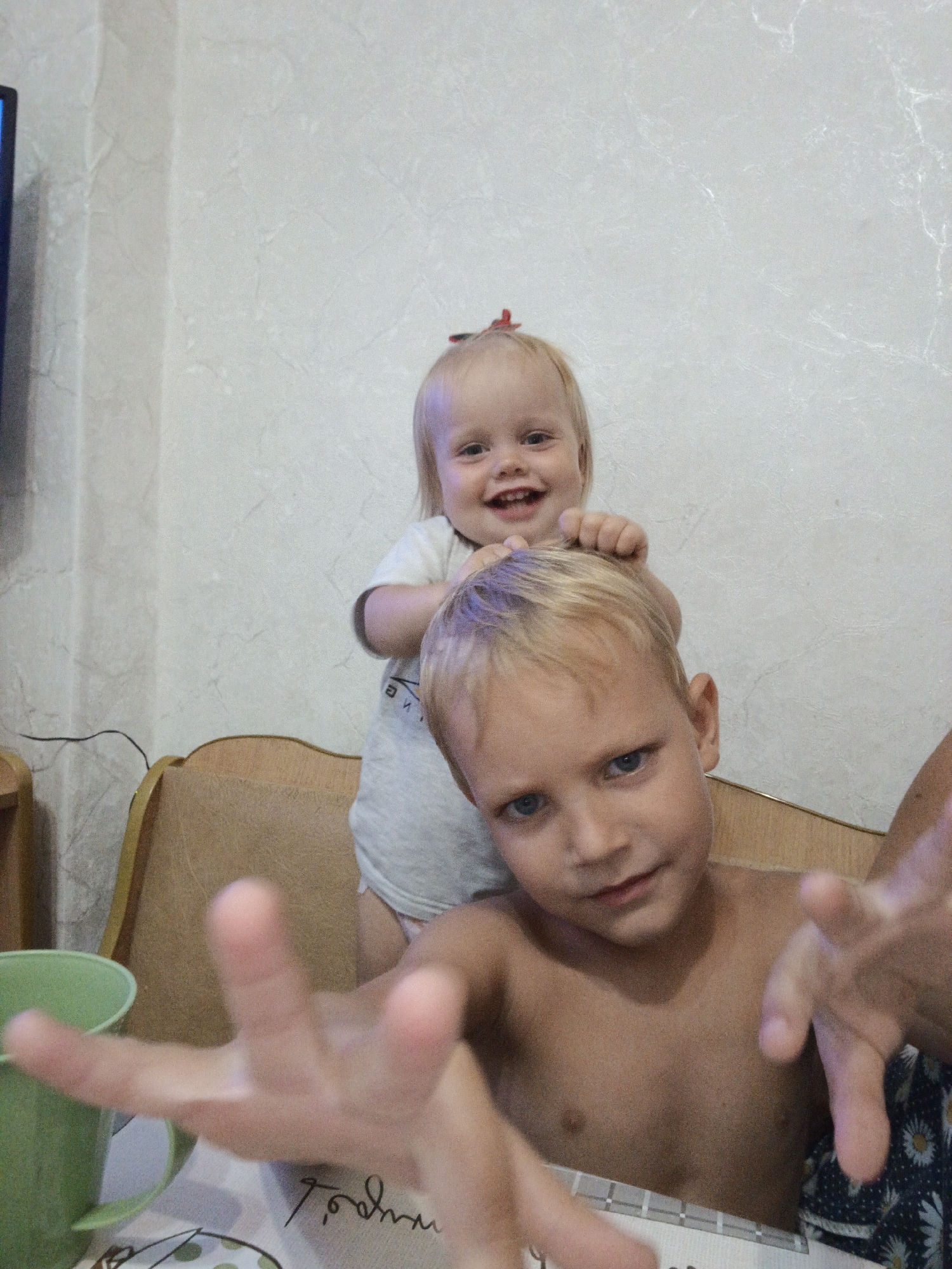 The media file shows two young children in an indoor setting, likely a home in Medvedyovka, Russia. The foreground is dominated by a young boy, shirtless and looking directly at the camera with a playful expression. His hands are extended towards the viewer, appearing to reach out. Behind him, perched on his head or shoulders, is a younger child, a baby or toddler, also smiling and looking towards the camera. This younger child has a small red bow in her hair. The background features a textured, light-colored wall and a portion of a chair or couch. A green cup is visible to the left of the frame, and a table with some patterned placemats or a tablecloth is in the lower part of the image. The lighting suggests it is daytime. The overall mood is one of playful interaction and candid family moment. There is some indistinct handwritten text on the table.