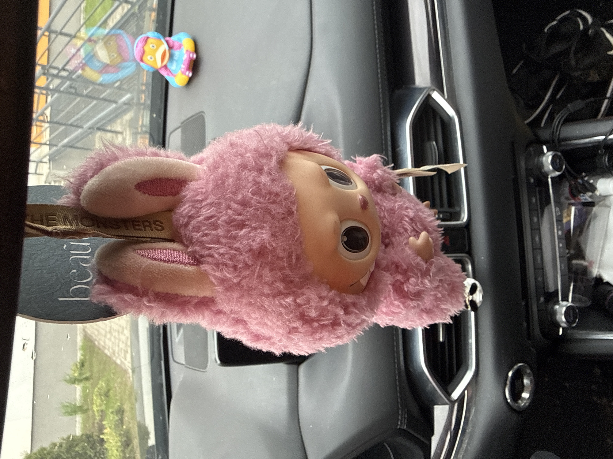 The image shows the interior of a vehicle, likely a car, with a pink, furry doll with a plastic face positioned on the dashboard. The doll has bunny-like ears with pink accents. A tag attached to the doll reads "THE MONSTERS" and "beaul". In the background, through the windshield, are reflections of buildings or structures. A small, colorful toy with a yellow face and blue attire is stuck to the upper left corner of the windshield. The dashboard is black and grey with metallic trim around the air vents. To the right, part of the car's console with buttons and a screen are visible. No people are visible in the image. It is impossible to determine if the image was taken in Berezyna, Ukraine, based on the visual information.