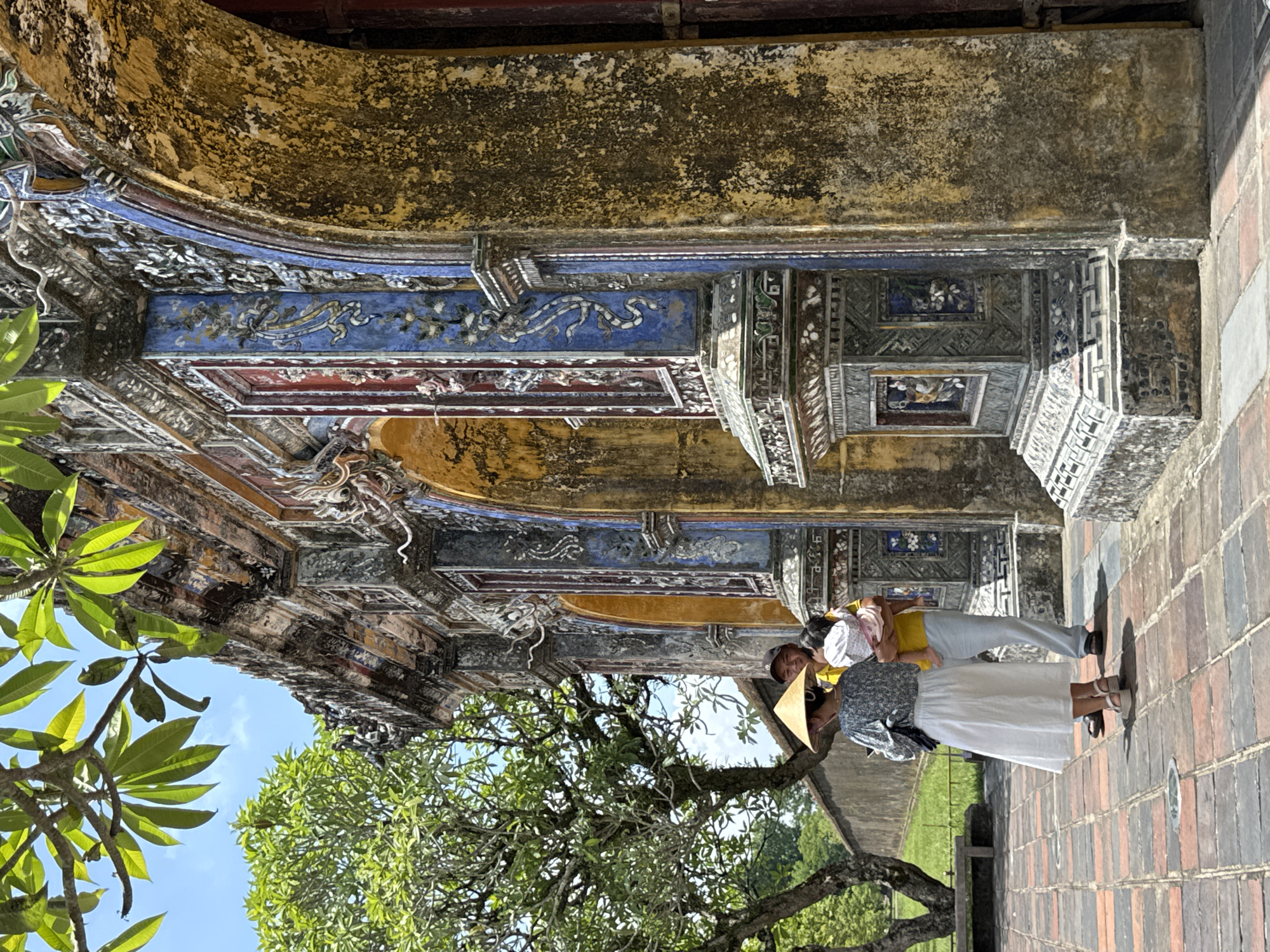 In Phú Xuân Ward, Vietnam, under a bright sky with leafy branches overhead, a family stands near a vibrantly decorated historic structure with arched entrances. The architecture is ornate, featuring yellow, blue, and red tiled embellishments with intricate patterns. A woman wearing a conical hat and a floral top with a white skirt faces away from the camera, looking towards the structure. Beside her, a man in a yellow shirt and light pants holds a baby dressed in pink. The ground is paved with multi-colored bricks. To the left, a lush green tree stands tall beside a stone wall and a weathered wooden bench. The scene captures a moment of cultural exploration and family connection within an ancient setting.