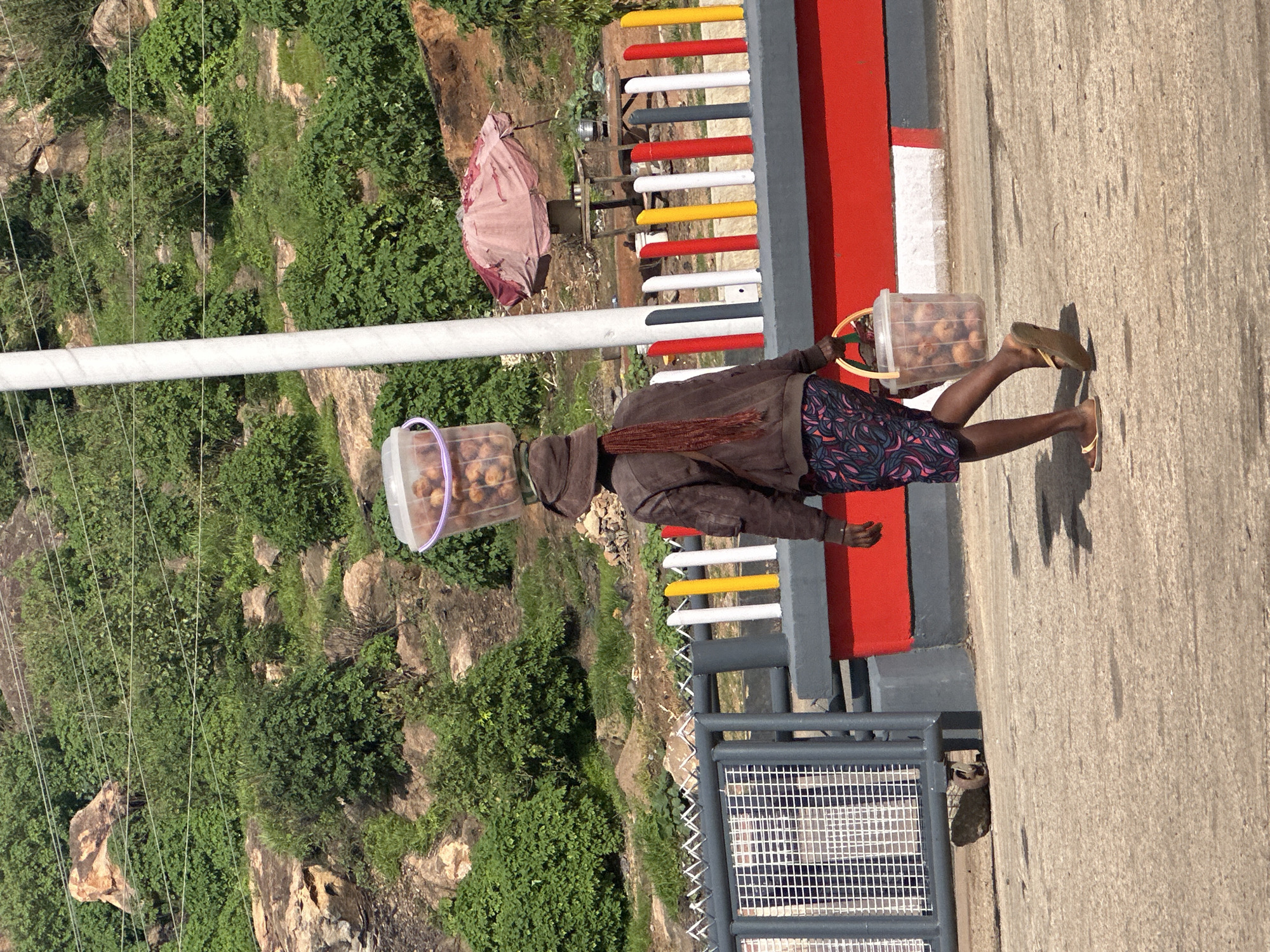 A person wearing a brown jacket, a hat, and patterned shorts walks on a paved surface, carrying a bucket of fruit on their head and another bucket in their hand. They are walking past a colorful fence with red, white, and yellow posts and a red and gray base. In the background, there is lush green vegetation and rocky terrain, characteristic of the landscape around Gura Top, Nigeria. A small, open-air stall with a pink umbrella and a table with cooking pots is visible further in the background. The overall scene depicts a person engaged in street vending in a natural, hilly environment.