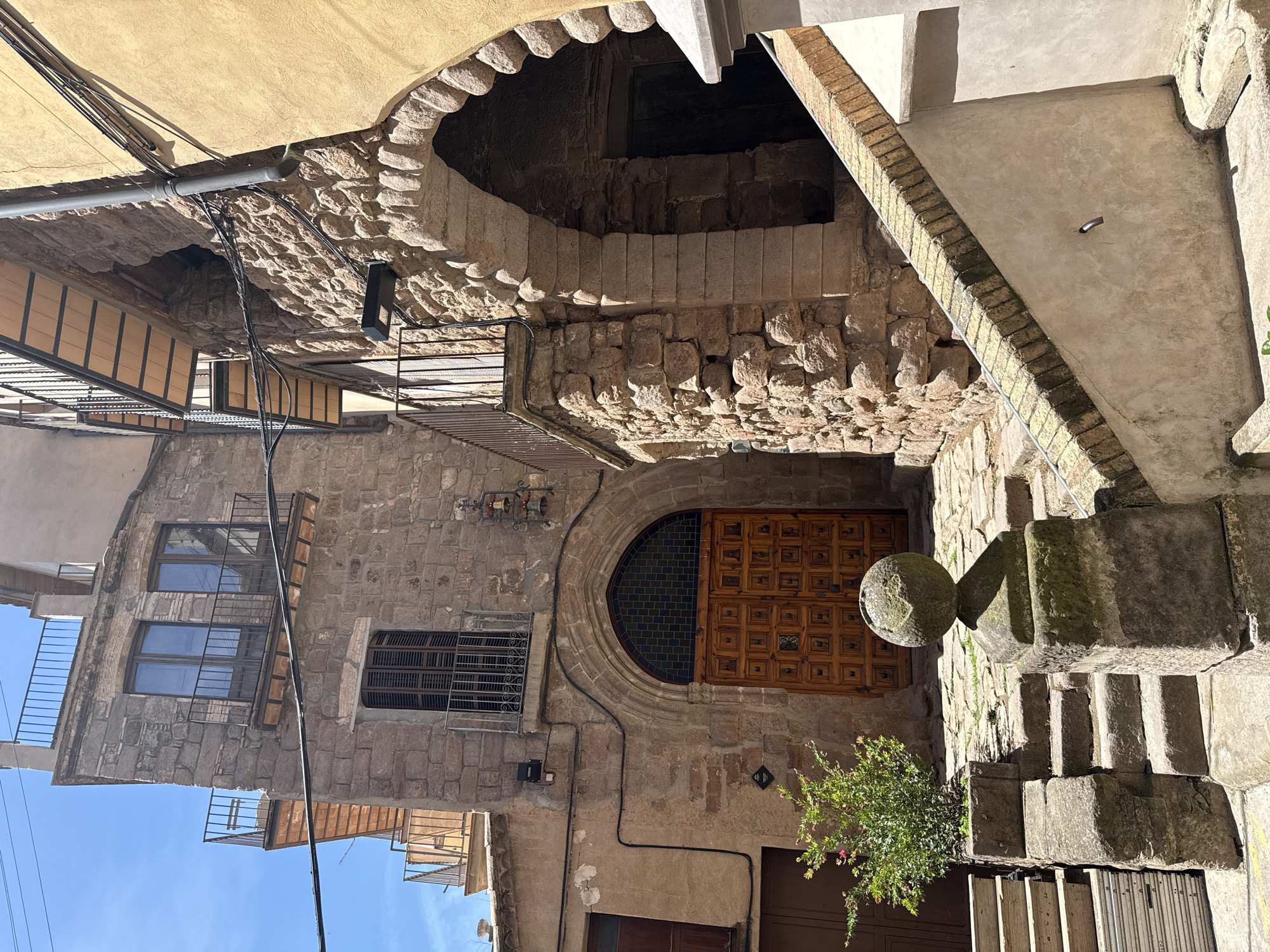The image captures a narrow, stone-paved alleyway in Cardona, Spain, leading towards a large, ornate wooden door set within a Gothic archway. The architecture is predominantly made of rough-hewn, reddish-brown stones, with contrasting lighter stone accents around the arched entrance. Balconies with wrought-iron railings jut out from the upper floors of the stone buildings. A bright yellow stucco building borders the alley on the right, featuring a similarly arched entryway. The foreground is dominated by a stone staircase with a weathered, spherical finial atop a stone pedestal. A small green bush with pink flowers is planted to the left of the stairs. The scene is bathed in bright sunlight, casting strong shadows. The sky is a clear blue, with a few thin power lines visible against it. There are no people or animals visible in the frame.