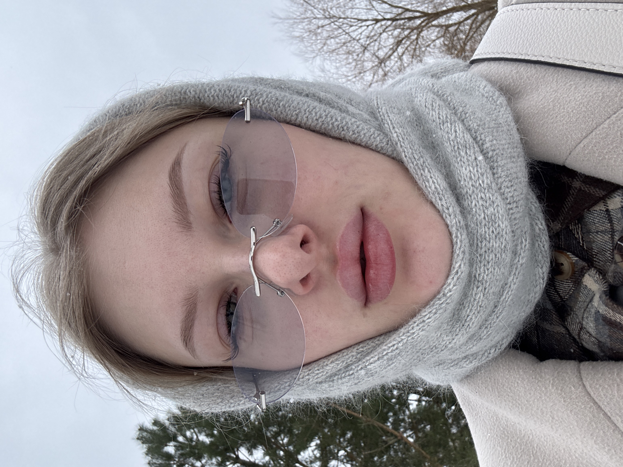 A close-up, rotated portrait image shows a young woman outdoors in a winter setting. She has blonde hair, visible beneath a light grey, ribbed knitted scarf that functions as a hood, covering most of her head and neck. She is wearing rimless, light-tinted oval sunglasses. Her full lips are slightly parted. Small white flakes, possibly snow or frost, are visible on her hair and scarf, indicating cold weather. She is also wearing a light-colored coat, possibly wool, with a contrasting strap from a handbag visible on her shoulder.
In the background, out of focus, are bare tree branches and the dark green needles of evergreen trees, characteristic of a winter landscape. The sky above is bright and overcast, suggesting daytime. The overall impression is a stylish pose in a chilly outdoor environment, consistent with winter conditions in Perm, Russia. No text is visible in the image.