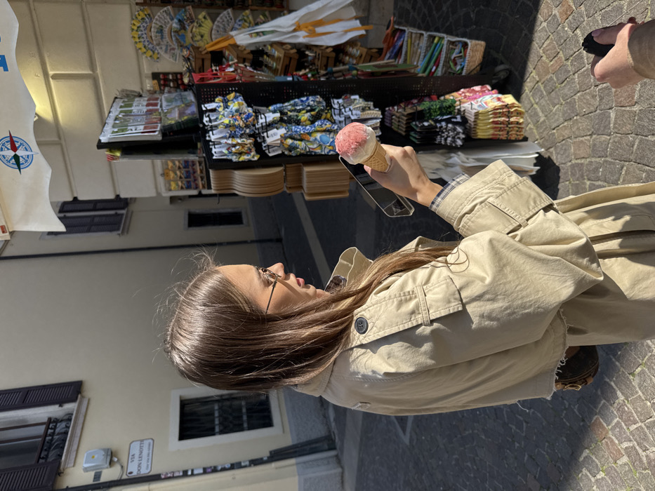 The image depicts an outdoor scene on Via Don Lenotti in Peschiera del Garda, Italy. A female subject with long brown hair, wearing a tan trench coat with plaid cuff and collar lining, and thin-rimmed eyeglasses, stands holding a single-scoop ice cream cone in her left hand. The ice cream is light pink with a white top layer. She looks slightly downwards and to her right. A small, dark handbag is partially visible on her right side, under her coat.
To the right of the female, a street vendor's display rack is visible. It features multiple shelves and hooks showcasing various merchandise, including brightly patterned swimwear (many with a lemon motif), postcards, and decorative fans. Above the stall, a white awning displays a red, blue, and white compass rose logo.
The background consists of light-colored stucco buildings with dark window shutters. A street sign affixed to the building on the left reads "VIA DON LENOTTI". The ground is paved with cobblestones. The scene is illuminated by direct sunlight, casting clear shadows. The partial right hand of a second individual, holding a black object, is visible in the lower right foreground.