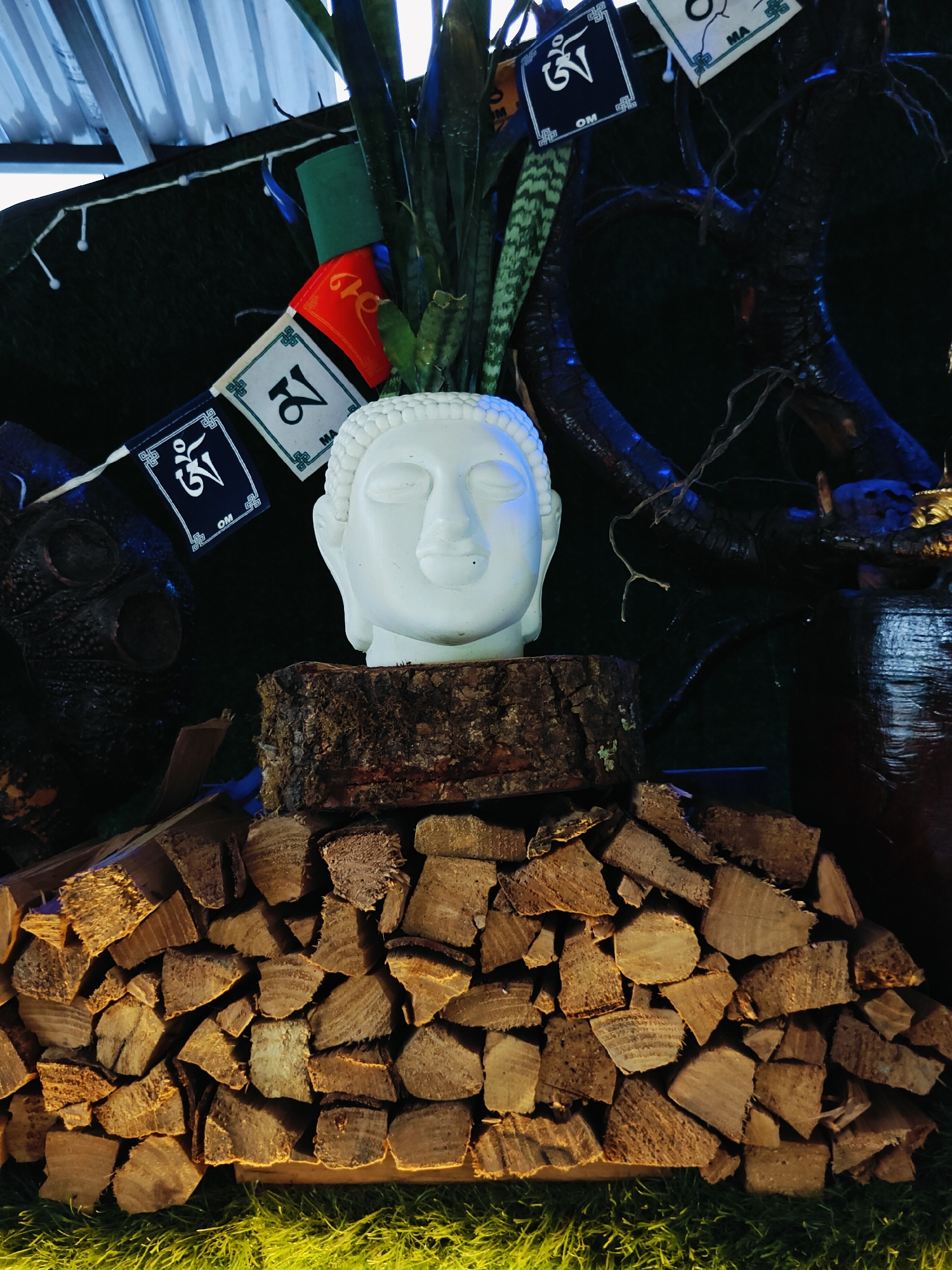 This image depicts an outdoor arrangement in Pokhara, Nepal, featuring a white Buddha head sculpture placed atop a circular wood log. The log rests on a neatly stacked pile of firewood, which is situated on bright green artificial grass. Behind the Buddha head, a snake plant is visible, and a string of prayer flags with Tibetan script adorns the background. The overall scene has a spiritual and decorative ambiance, possibly set in a garden or courtyard.