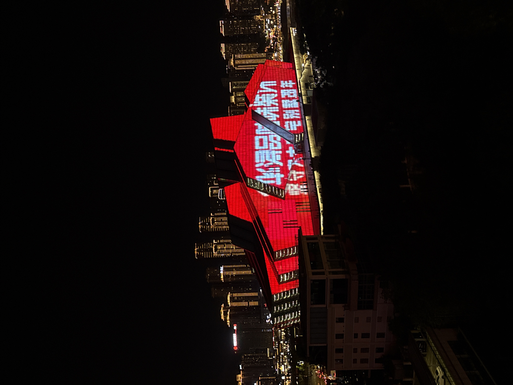 A wide-angle, elevated night view captures a brightly lit, modern architectural structure in the foreground, dominating the frame with its vibrant red facade adorned with white LED text. The building, which appears to be a performance venue or cultural center, features angular designs and is illuminated from within and on its exterior. The LED text, displayed in Chinese characters, is large and prominent, suggesting a special event or announcement.
Behind this main structure, a cityscape unfolds, characterized by numerous high-rise buildings in the background, many of which are also illuminated with warm, yellow lights. These buildings indicate a dense urban environment. The overall scene is set at night, with a dark sky and a sense of depth created by the layered lights.
The location is identified as 江北城街道, China. The image is taken from a vantage point that overlooks the main building and the surrounding city, suggesting it might be from a hotel, another building, or a high vantage point within the urban area. There are no discernible people or distinct actions captured in the immediate foreground of the main building, though the lights on the ground level of the building suggest activity. The image appears to be a photograph taken with a mobile device, given the resolution and slight graininess.