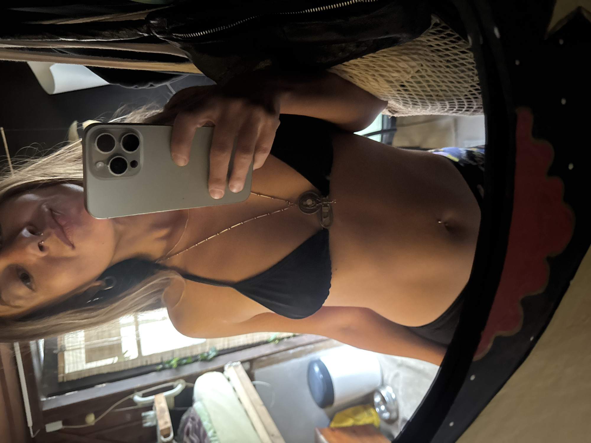 A woman with long brown hair is taking a selfie in a mirror. She is wearing a black bikini top and black bottoms. She has a nose ring and a belly button piercing. A silver necklace with a pendant is visible around her neck. She is holding a silver smartphone in her right hand, which is partially obscuring her chest. In the background, a bed with white bedding is visible on a shelf. To the left of the bed is a white cylindrical object with a gray lid. A metal bowl is on the floor below the shelf. To the right of the woman, a black jacket is hanging, and a cream-colored mesh bag is visible. The mirror has a black frame with red and white decorative elements. The overall environment appears to be an indoor space, possibly a room with wooden walls and a window with blinds.