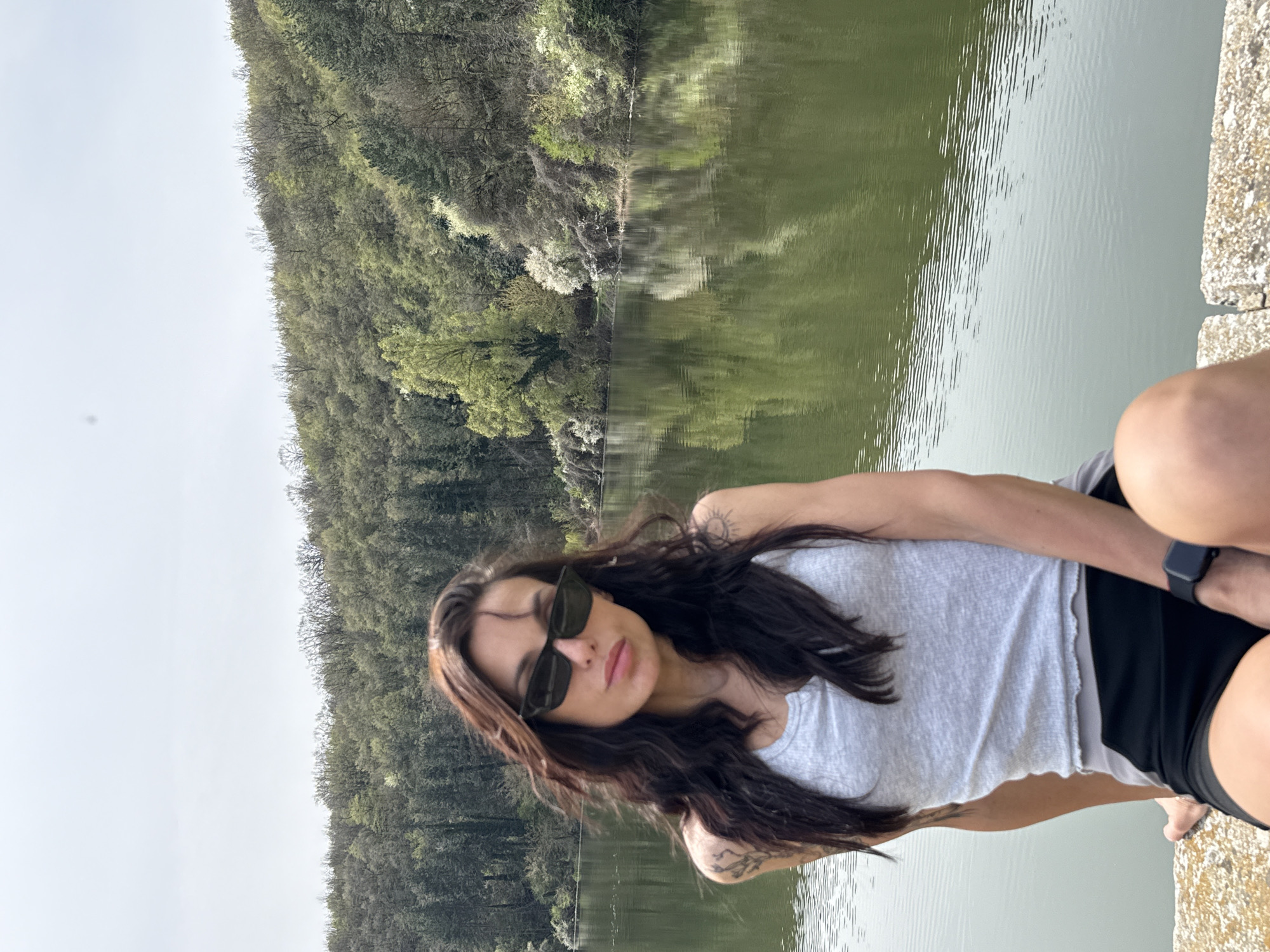 A woman is seated outdoors, near water. She wears sunglasses, a light-colored tank top, and dark shorts. Her dark hair is long and loose. A watch is visible on her wrist. The background features a lake or pond bordered by trees and foliage. It is daytime. While it is not possible to independently confirm the location as Pleven, Bulgaria, based on the visual information in this image, the environment and composition of the scene suggest a lakeside or park setting.