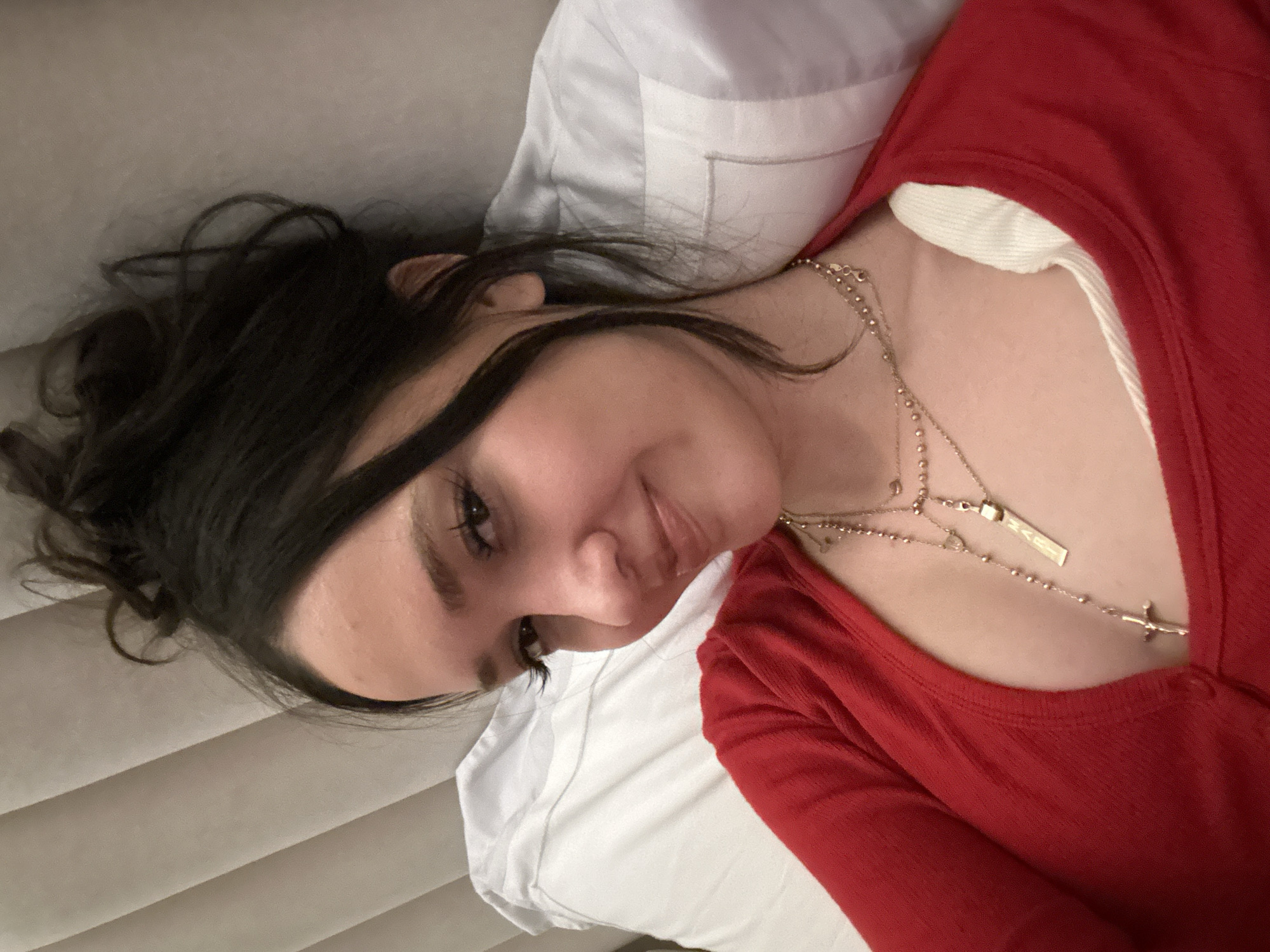 A young woman is depicted in a close-up, vertical shot, appearing to be lying down and looking towards the camera with a soft, relaxed smile. She has dark, long hair, styled loosely with some strands falling around her face. Her eyes are open, and she wears subtle eye makeup.
She is dressed in a red, long-sleeved top, worn over a white inner layer, the collar of which is visible at her neckline. Around her neck, she wears several layered gold-toned necklaces. One prominent necklace features a small, simple cross pendant. Another necklace has a rectangular bar pendant, oriented vertically, on which the word "HAITI" is clearly visible in capital letters. A third necklace consists of small gold beads.
Behind her head and upper body, a white pillow is visible, suggesting she is in a bed or on a cushioned surface. To the left side of the frame (her right), a light-colored, textured background with horizontal linear patterns can be seen, consistent with a padded headboard or wall paneling. The lighting is soft and even, highlighting her features. The overall impression is a serene and personal moment captured indoors. No specific cues for Dubai, United Arab Emirates, are visible in the image itself.